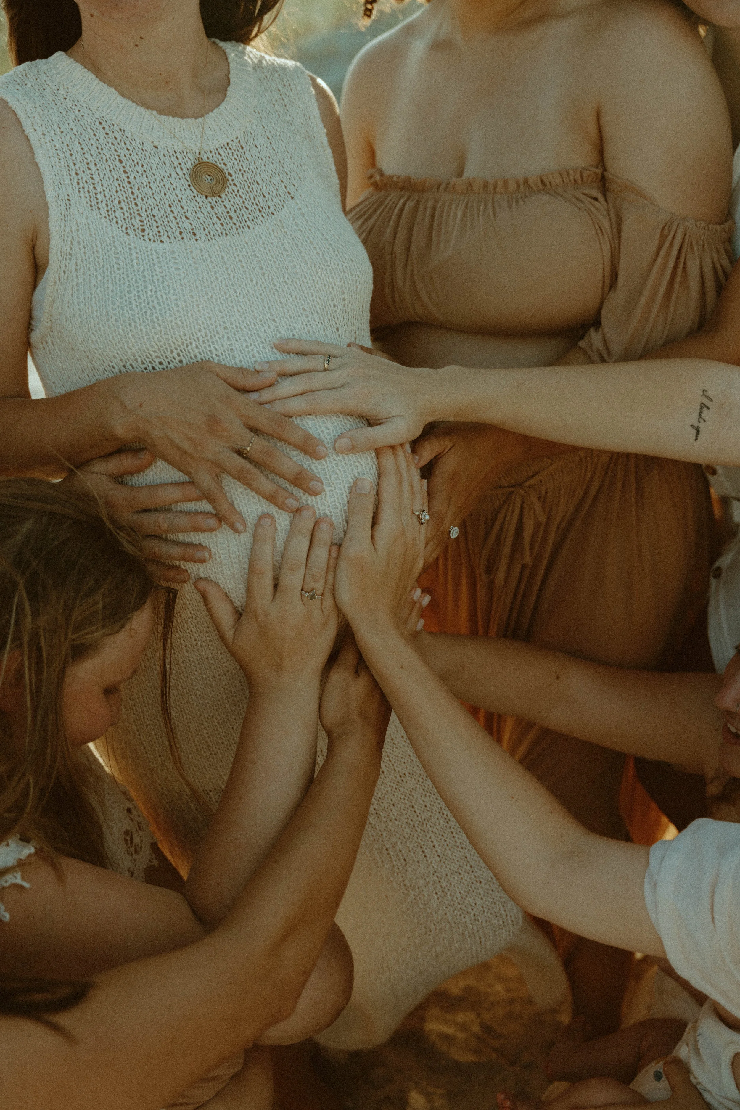 Multiple women and children touching and holding a pregnant woman’s belly during a celebration or gathering outdoors.