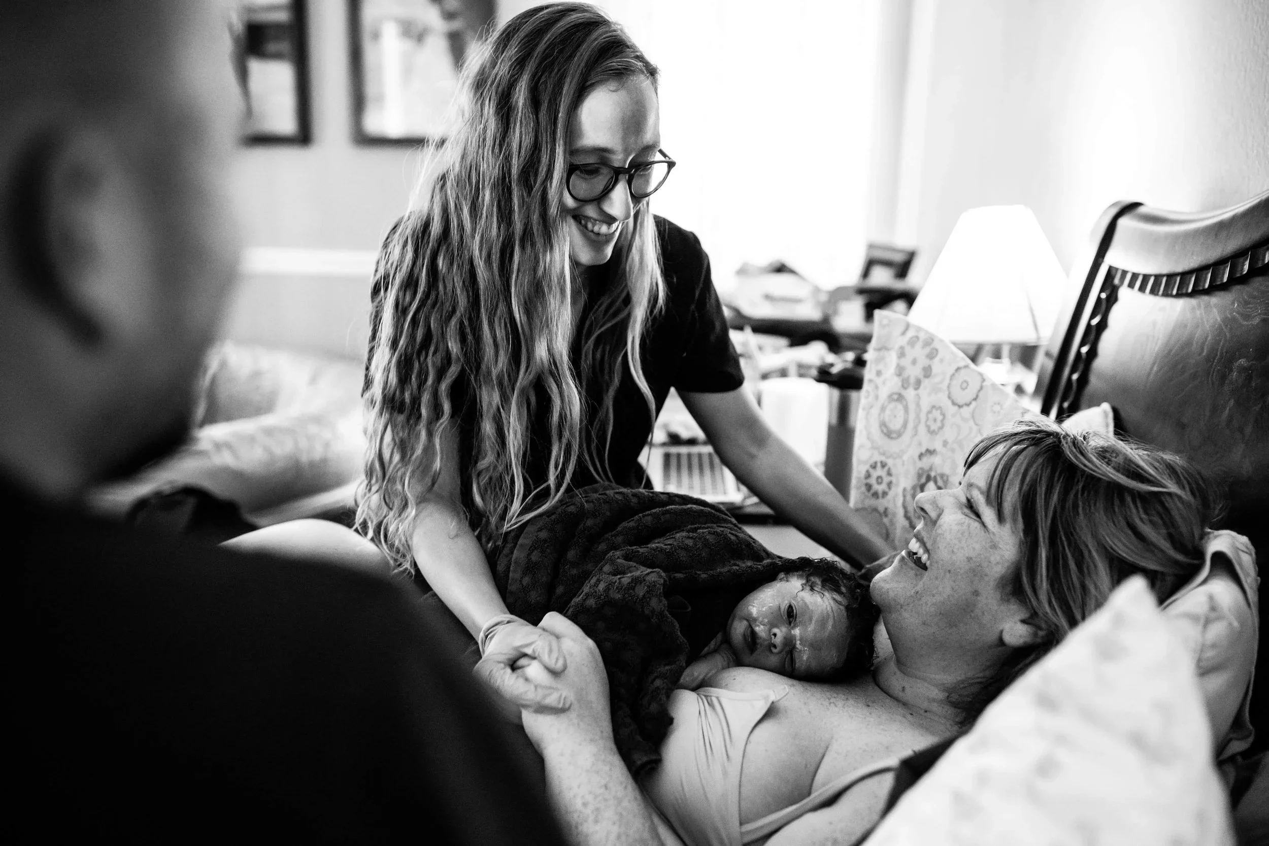 A woman lying in bed holding a newborn baby, surrounded by a smiling woman and another person in a hospital room.