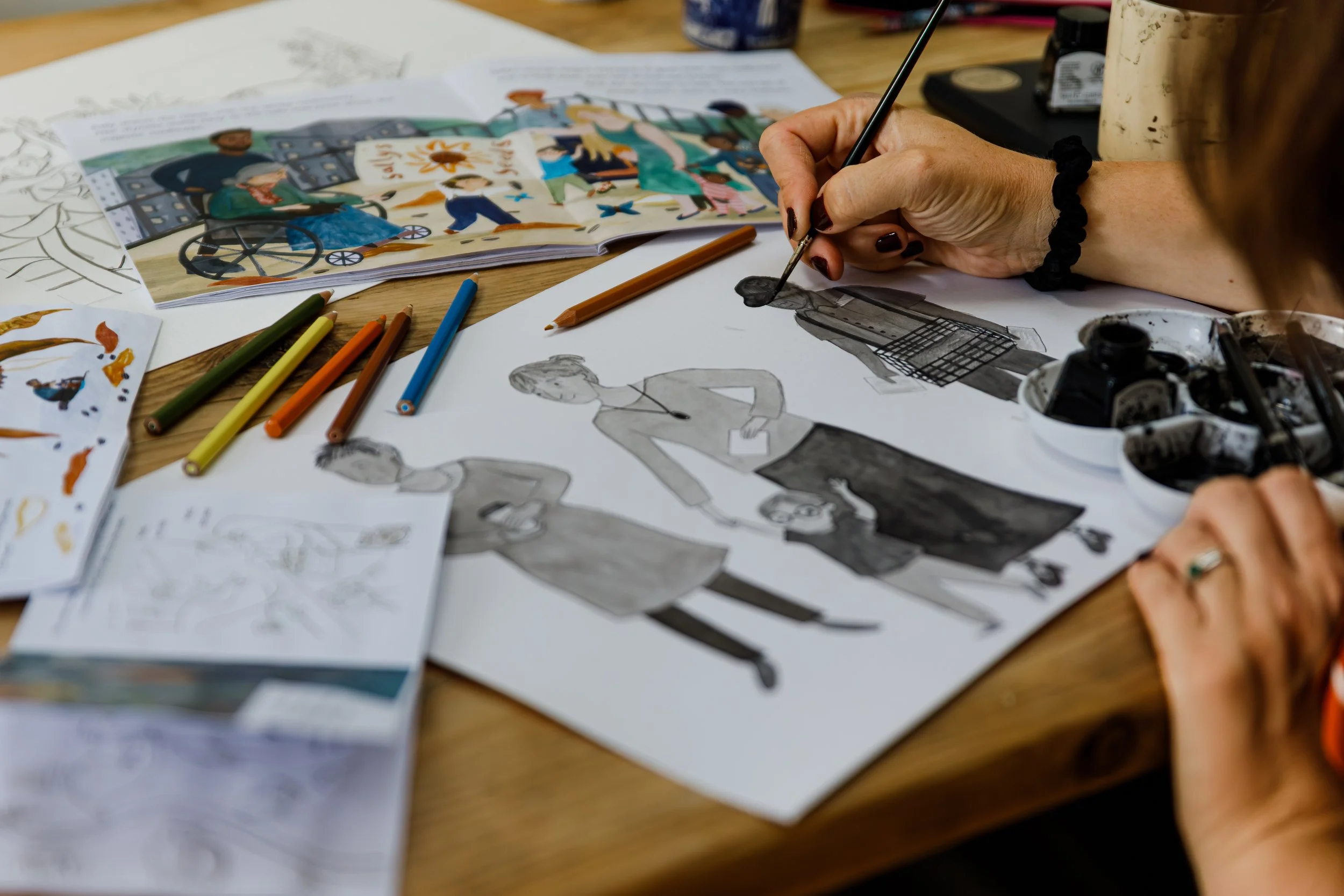 Person drawing a fashion figure on paper with colored pencils, surrounded by illustration sheets and art supplies on a wooden table.