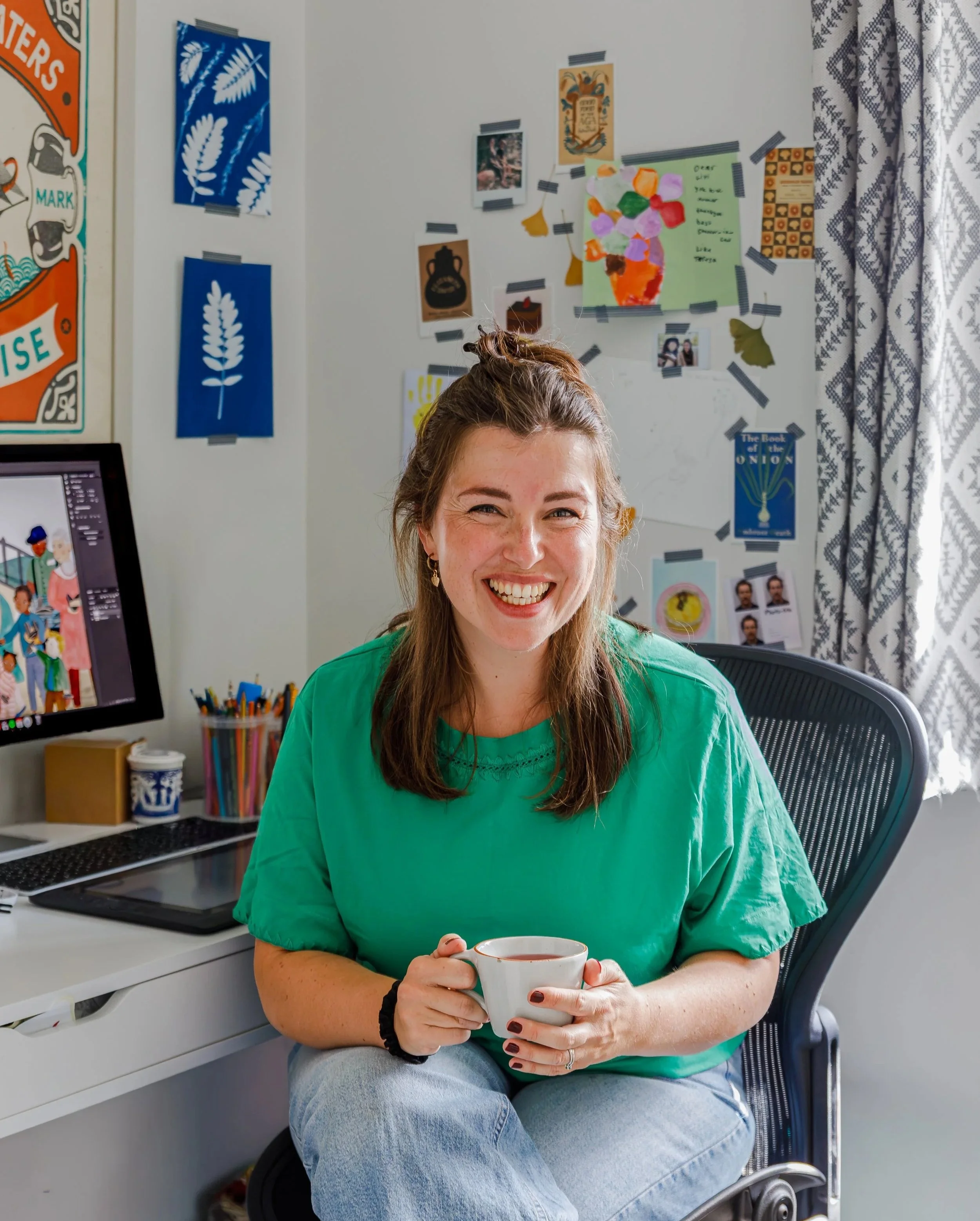 A woman with long brown hair wearing a green shirt and blue jeans is sitting on a black chair, smiling and holding a white mug. She is in a room with a white wall covered in colorful artwork and photos, a desk with a computer monitor, and a container of colored pens.
