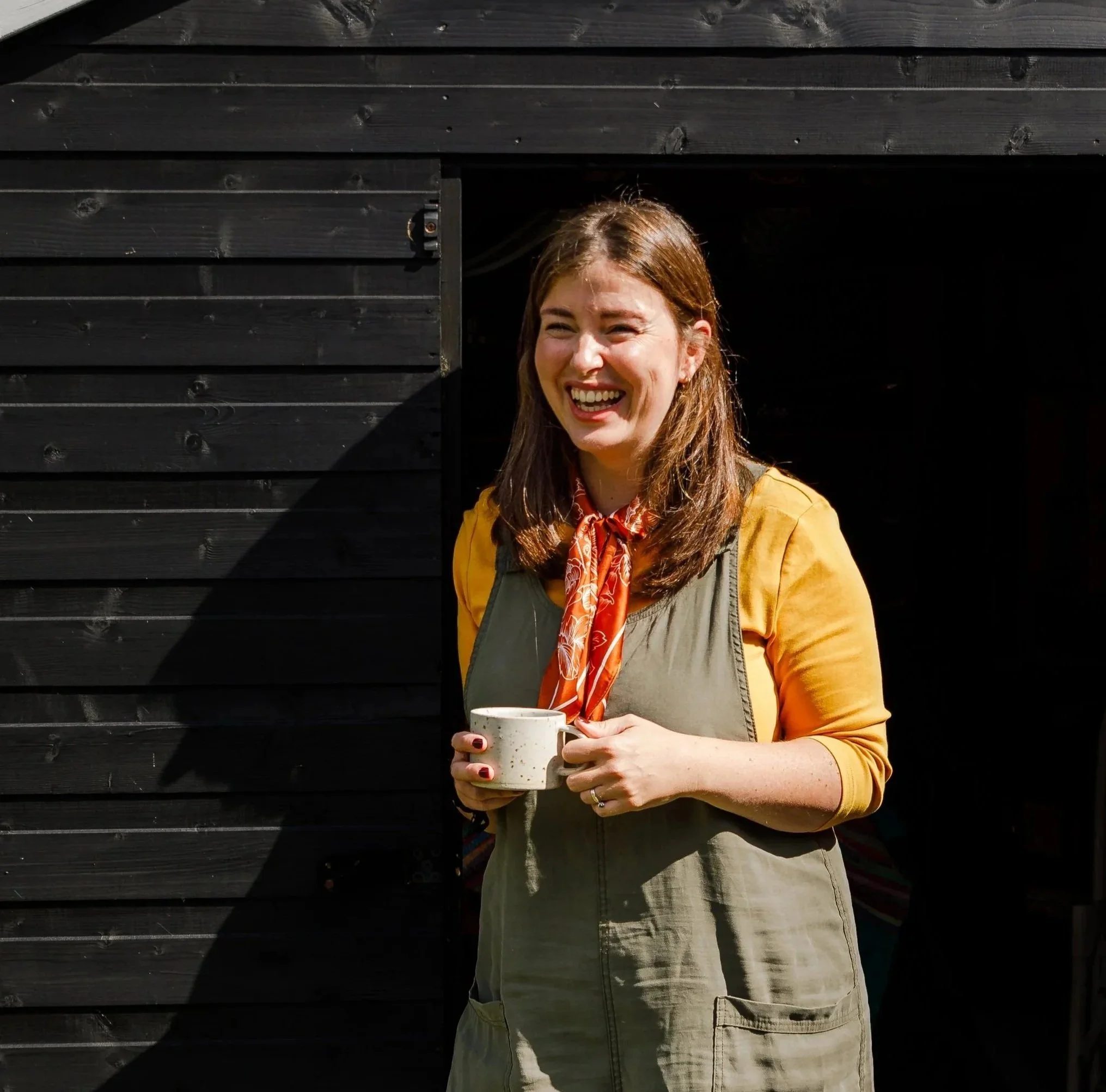 A woman with brown hair smiling and holding a mug, standing outside a black wooden building.