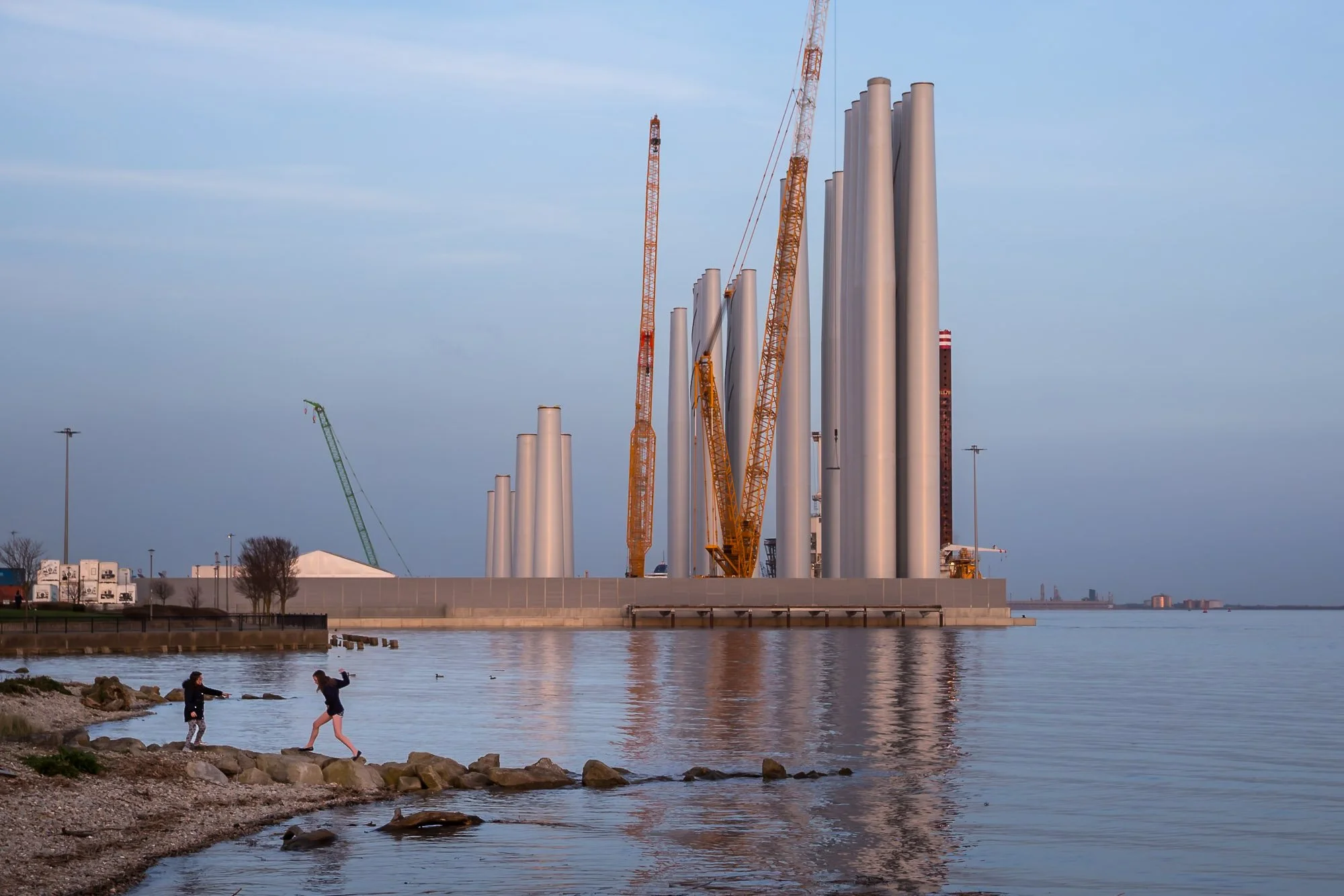 Two local girls play on the banks of the Humber with the turbine towers of Siemens Gamesa's factory on the former coal dock reflecting the late afternoon sunshine looming in the background