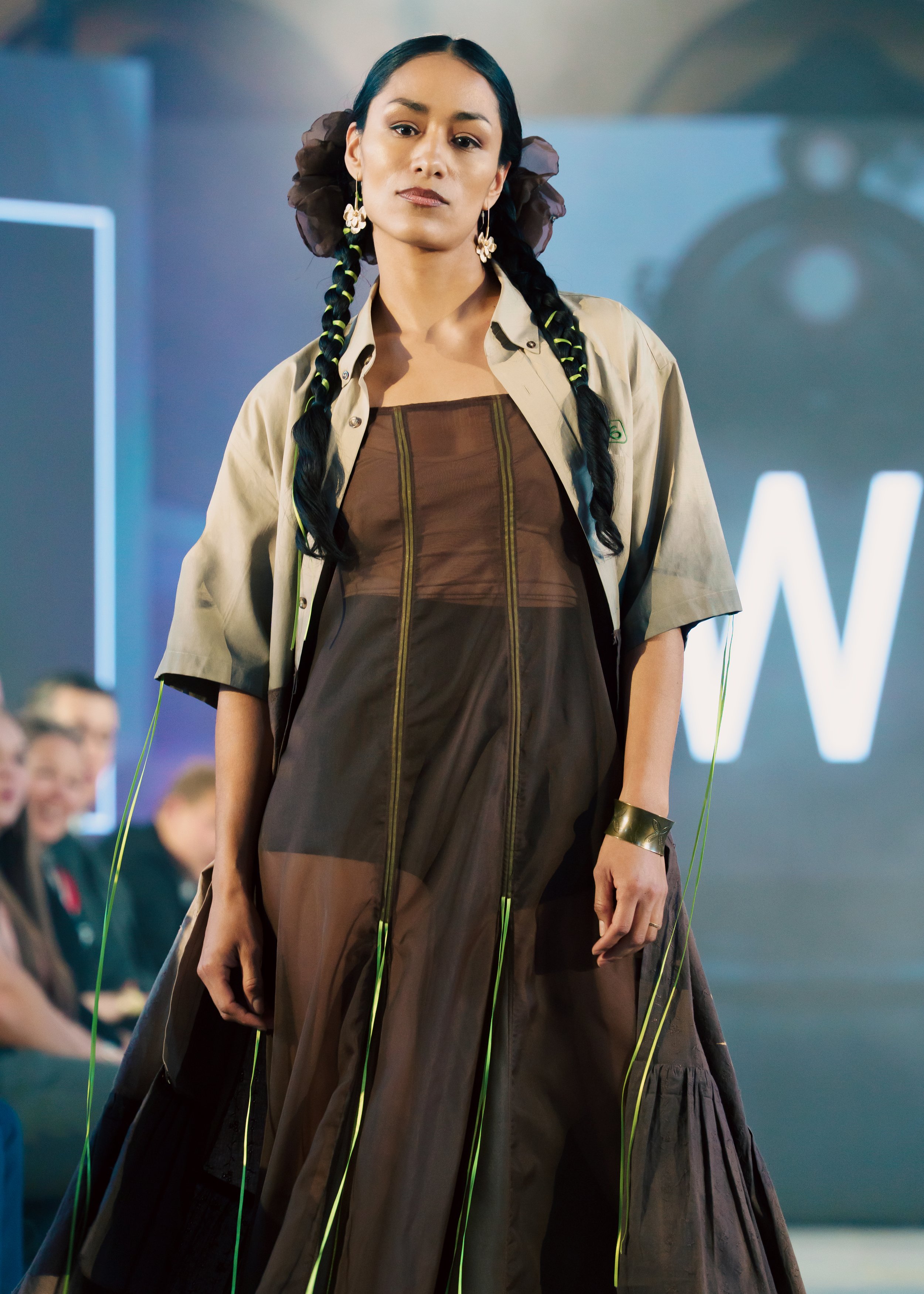 A fashion model with long black braided hair wearing floral earrings, a beige shirt, and a layered brown and black dress, walking on a runway during a fashion show.