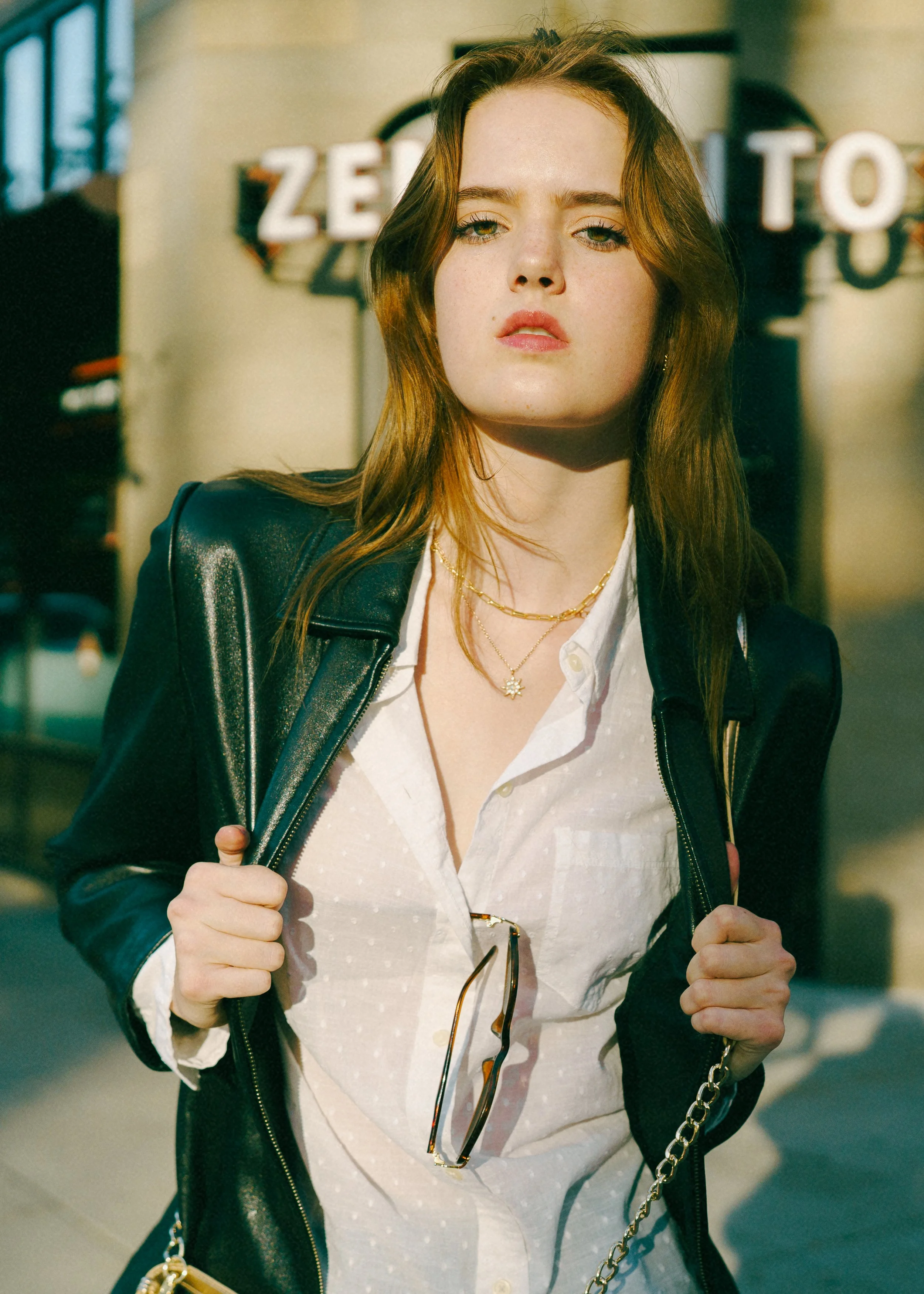 A fashion model with red hair and freckles wears a black leather jacket over a white shirt, posing for a photo while holding the straps. She has layered gold necklaces, including one with a star pendant, and sunglasses hanging from her shirt. 