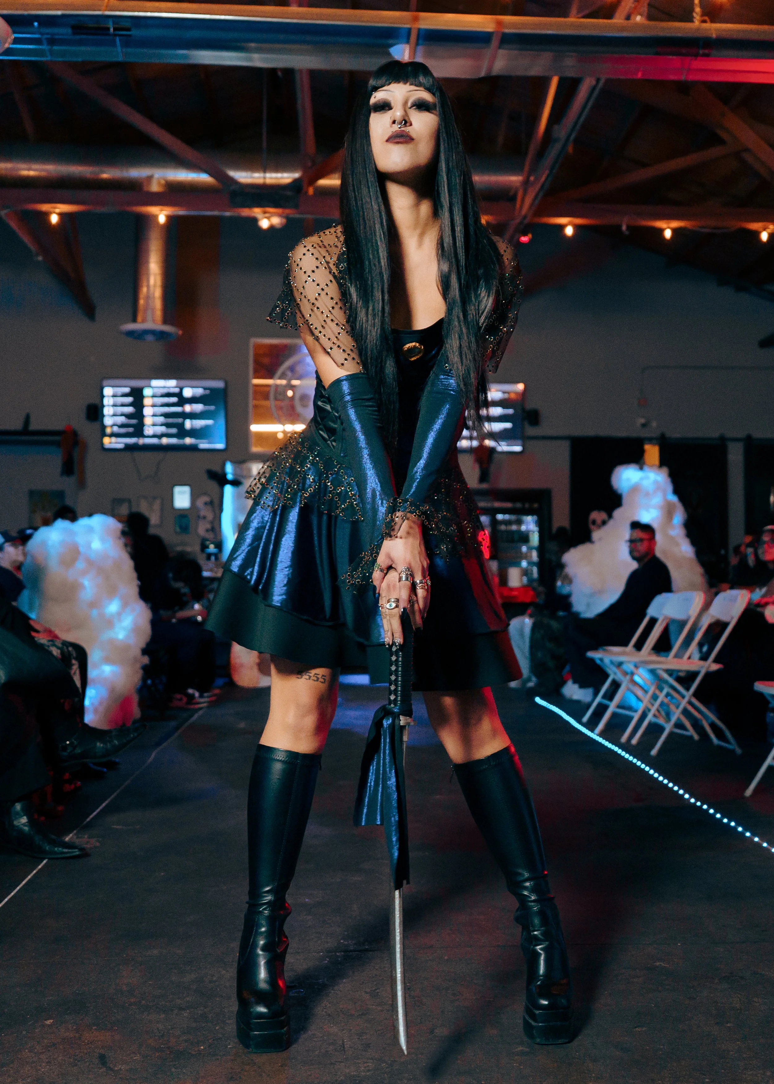 A fashion model with long black hair, dressed in a black shiny outfit with long gloves, standing in a fashion runway with string lights, people sitting on chairs, and smoke clouds in the background.