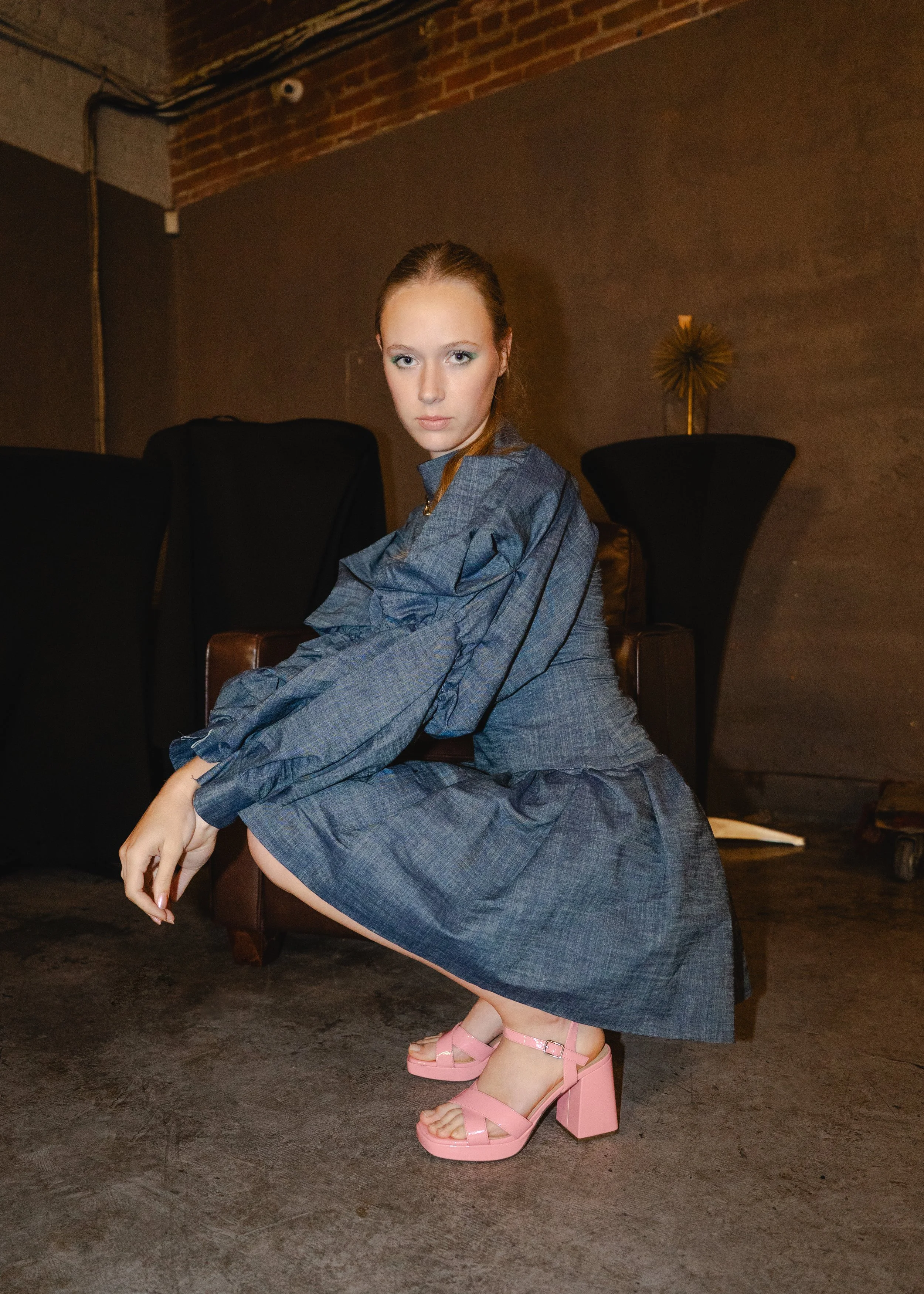 A fashion model with red hair wearing a blue checkered dress and pink high-heeled sandals squatting on a concrete floor in a dimly lit industrial-style room during a photo shoot.