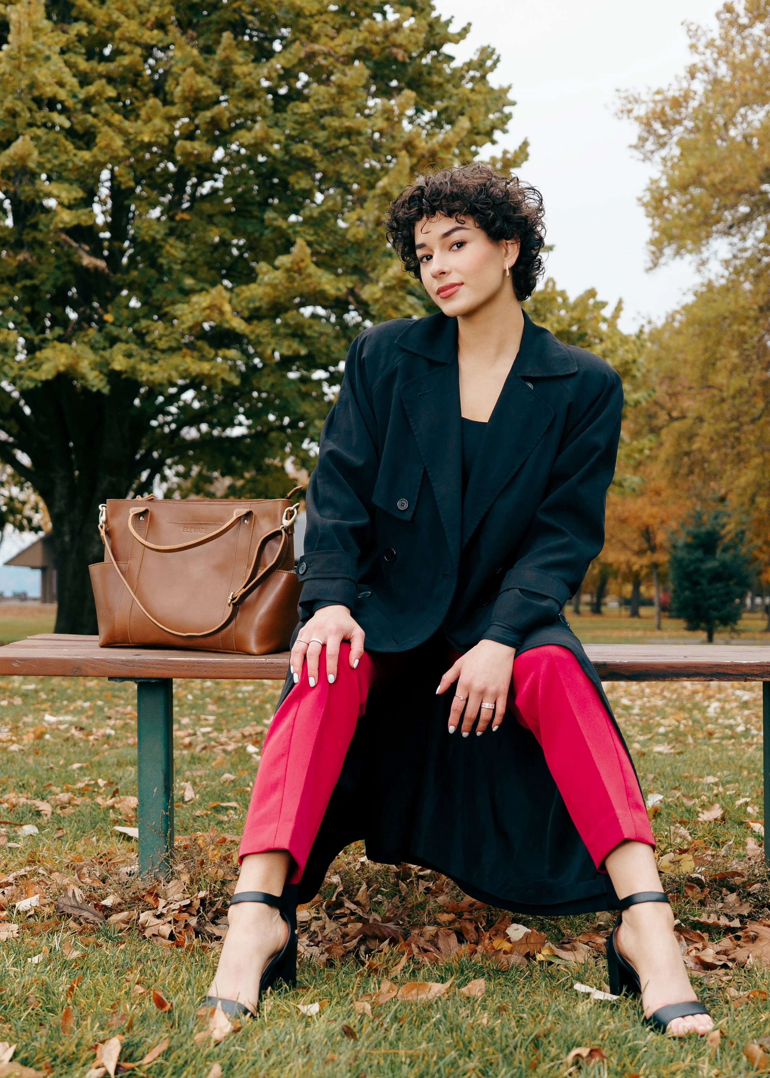 A fashion model with curly dark hair and earrings sitting on a park bench during autumn photography session, wearing a black jacket and red pants with black shoes, with a brown handbag beside her looking fabulous with a great smile. She is a fashion 