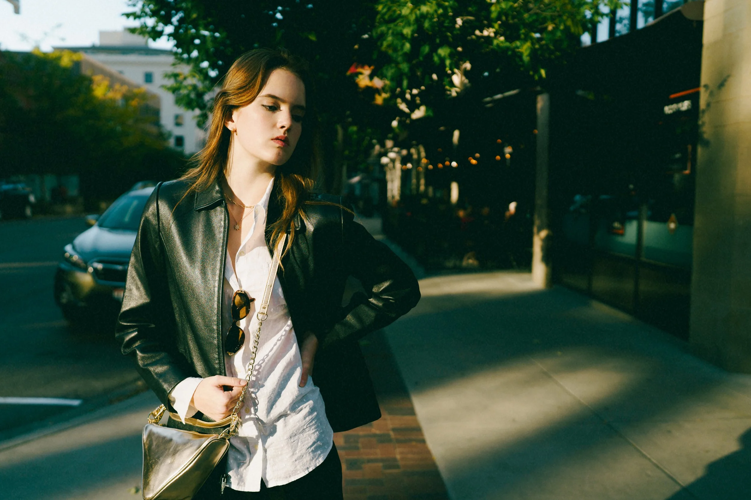 Young fashion model with long hair wearing a black leather jacket and white shirt, standing on a city sidewalk during dusk for a fashion shoot, holding a gold purse with sunglasses hanging from her shirt, looking down with a serious expression.