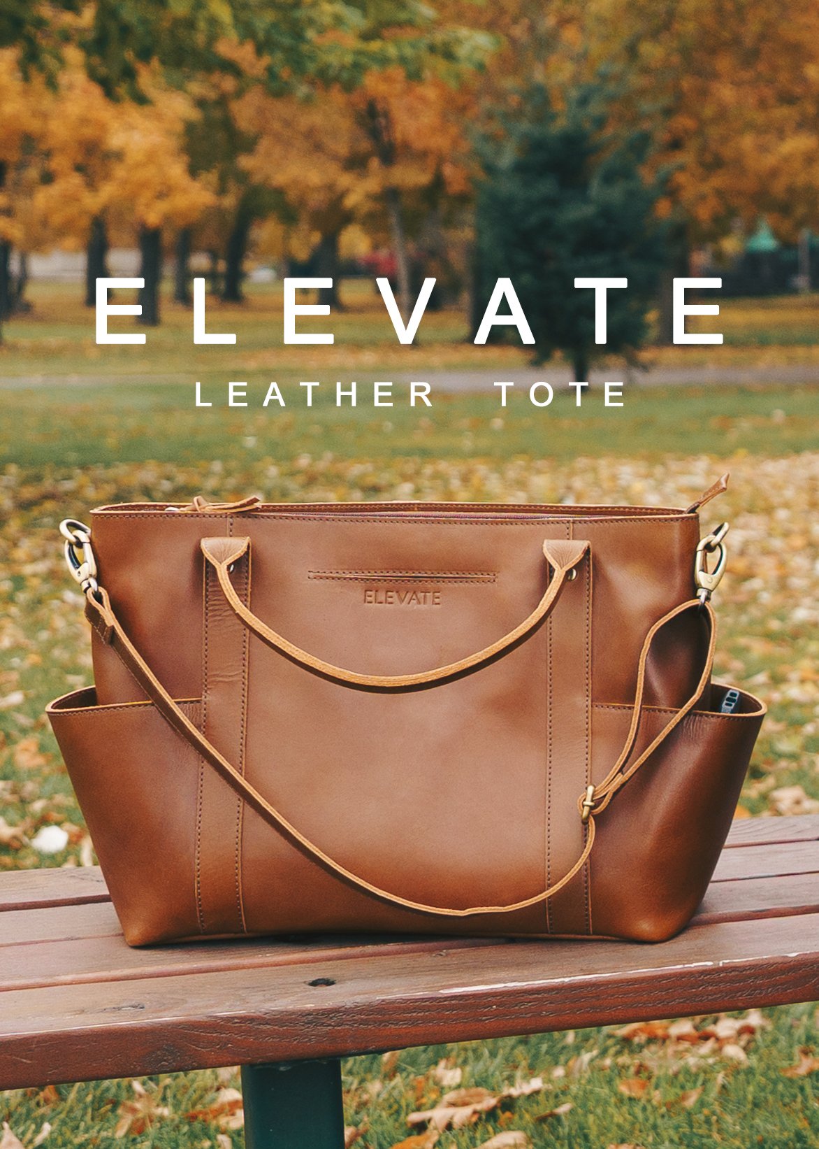 A brown leather tote bag with two handles and side pockets, displayed for a fashion shoot on a park bench with fall foliage in the background.