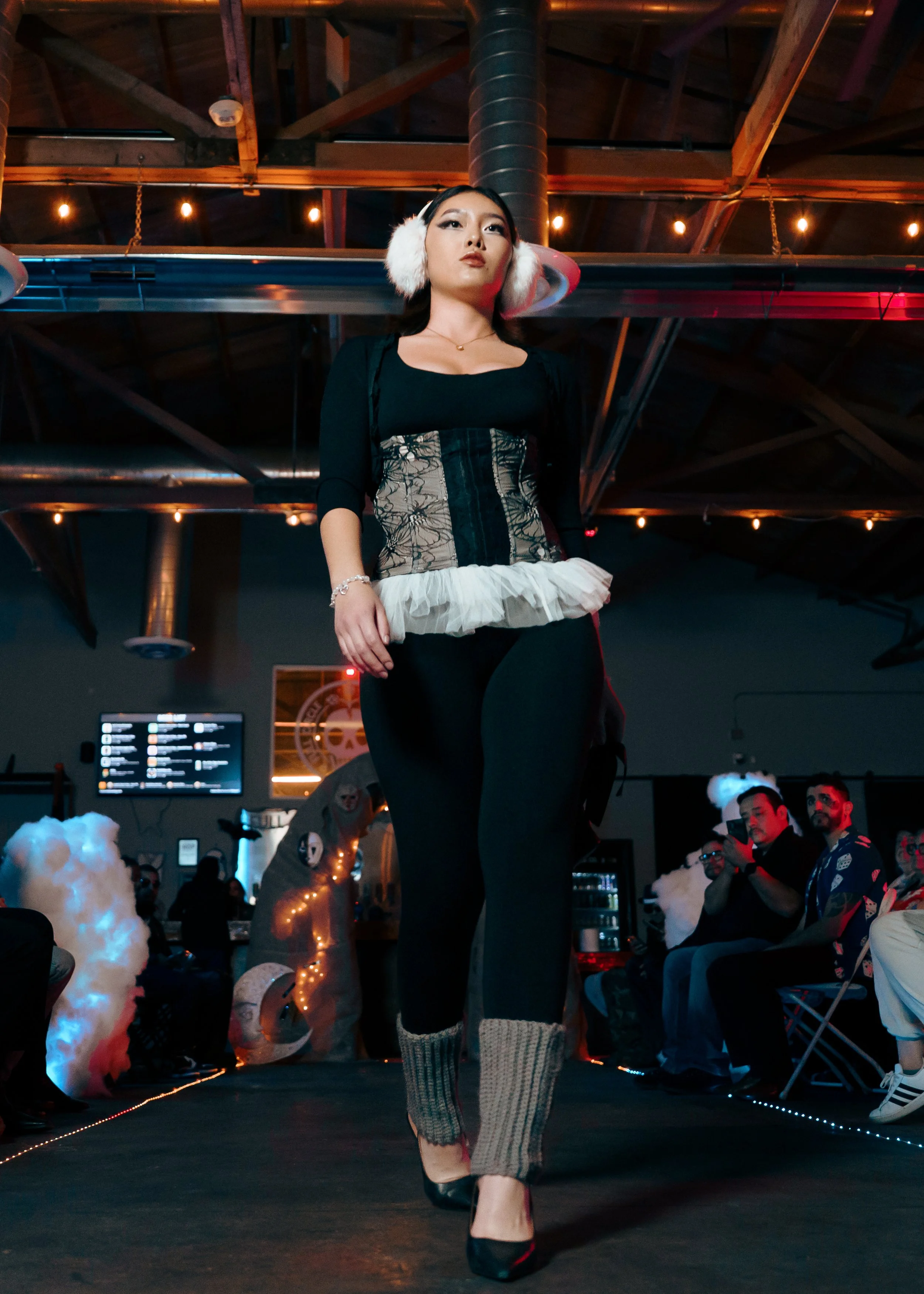 A fashion model is walking down a runway during a fashion show, wearing headphones, a black top, a corset with lace overlay and ruffled trim, black pants, and knitted leg warmers, with an industrial-style ceiling and audience in the background.
