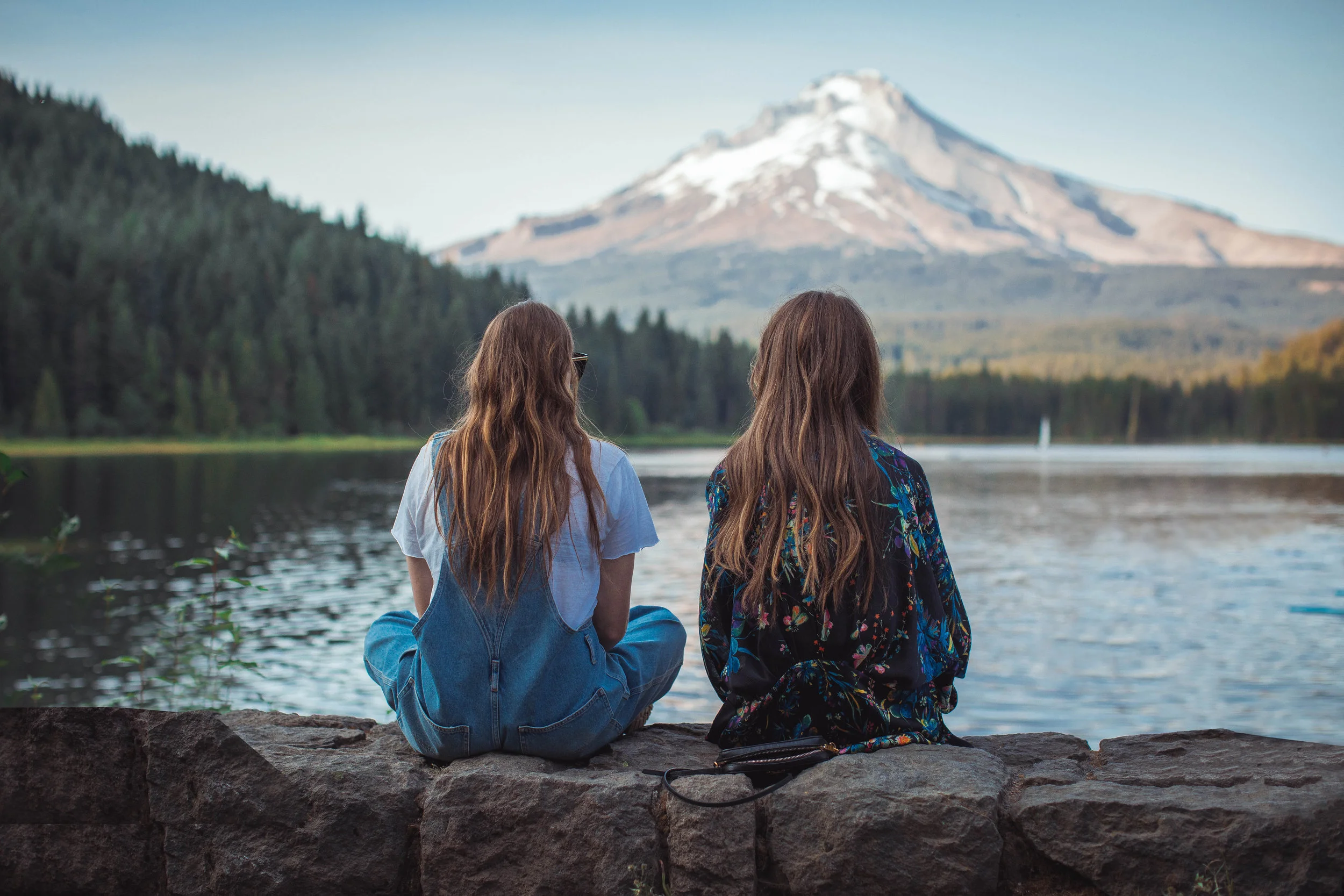 Two women enjoy a mountain view while sitting together lakeside. Enjoy life with emotional freedom, relieve stress, increase self confidence.
