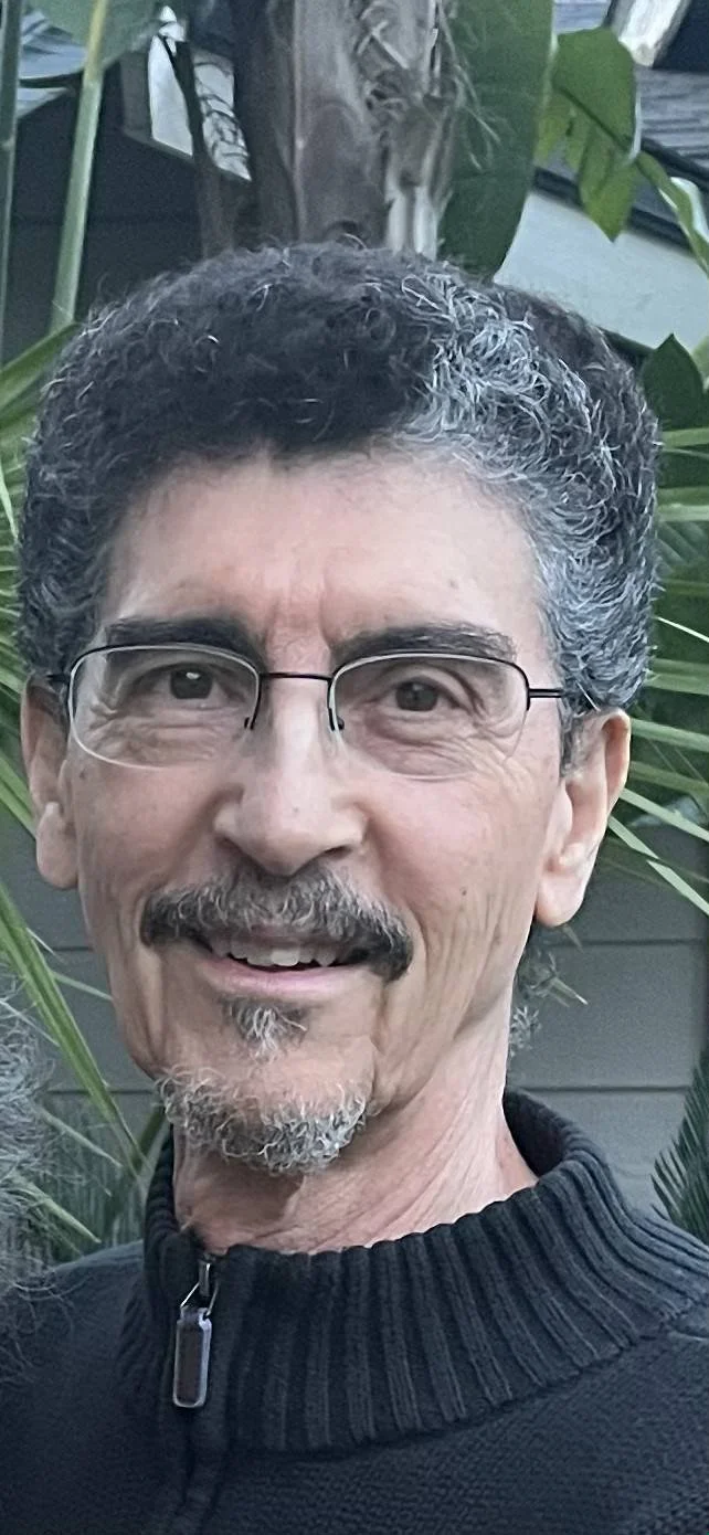 A middle-aged man with curly gray hair, glasses, and a goatee smiling outdoors, surrounded by green plants.