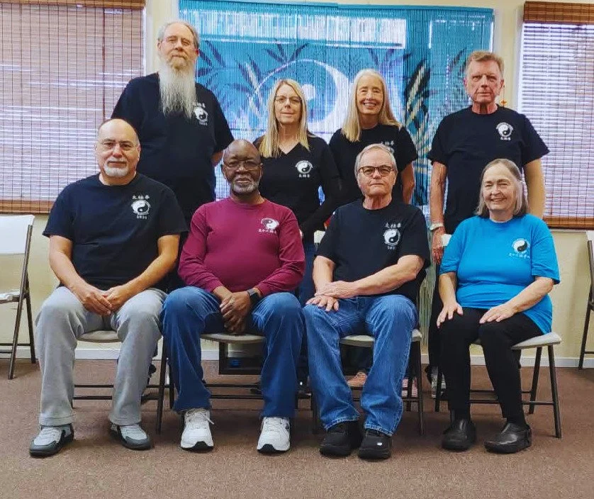 Group of nine diverse adults, some wearing matching t-shirts with a circular logo, posing indoors in front of a colorful mural with trees and leaves. They are seated and standing, smiling for the photo in a casual setting.