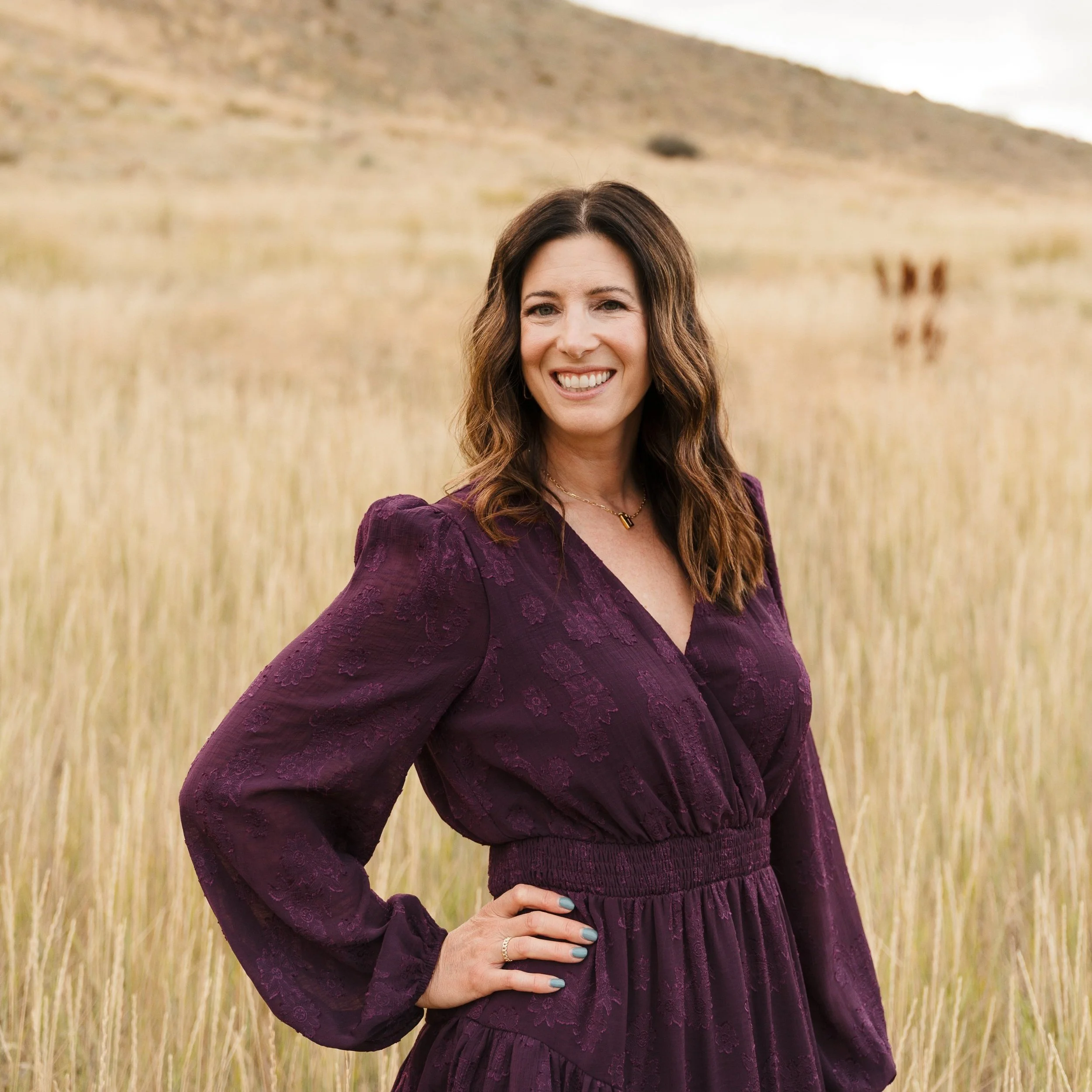 A woman with dark, wavy hair smiling, standing in a field of tall grass with hills in the background, wearing a purple dress.