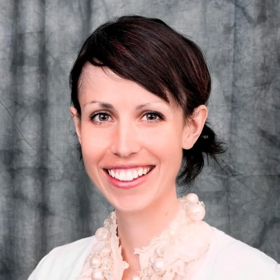 A smiling woman with short dark hair and light skin, wearing a white blouse with decorative pearls around the collar, standing in front of a gray textured background.