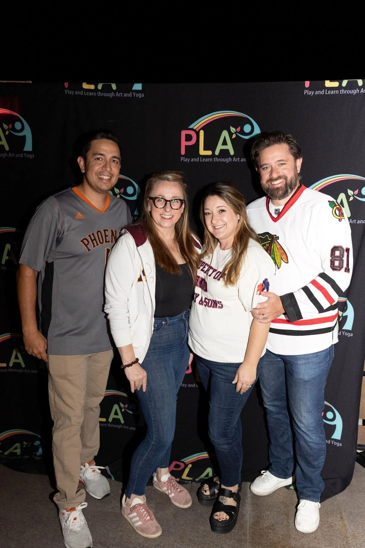 Four adults are standing together and smiling for a photo in front of a black backdrop with the Play and Learn through Art and Yoga logo.