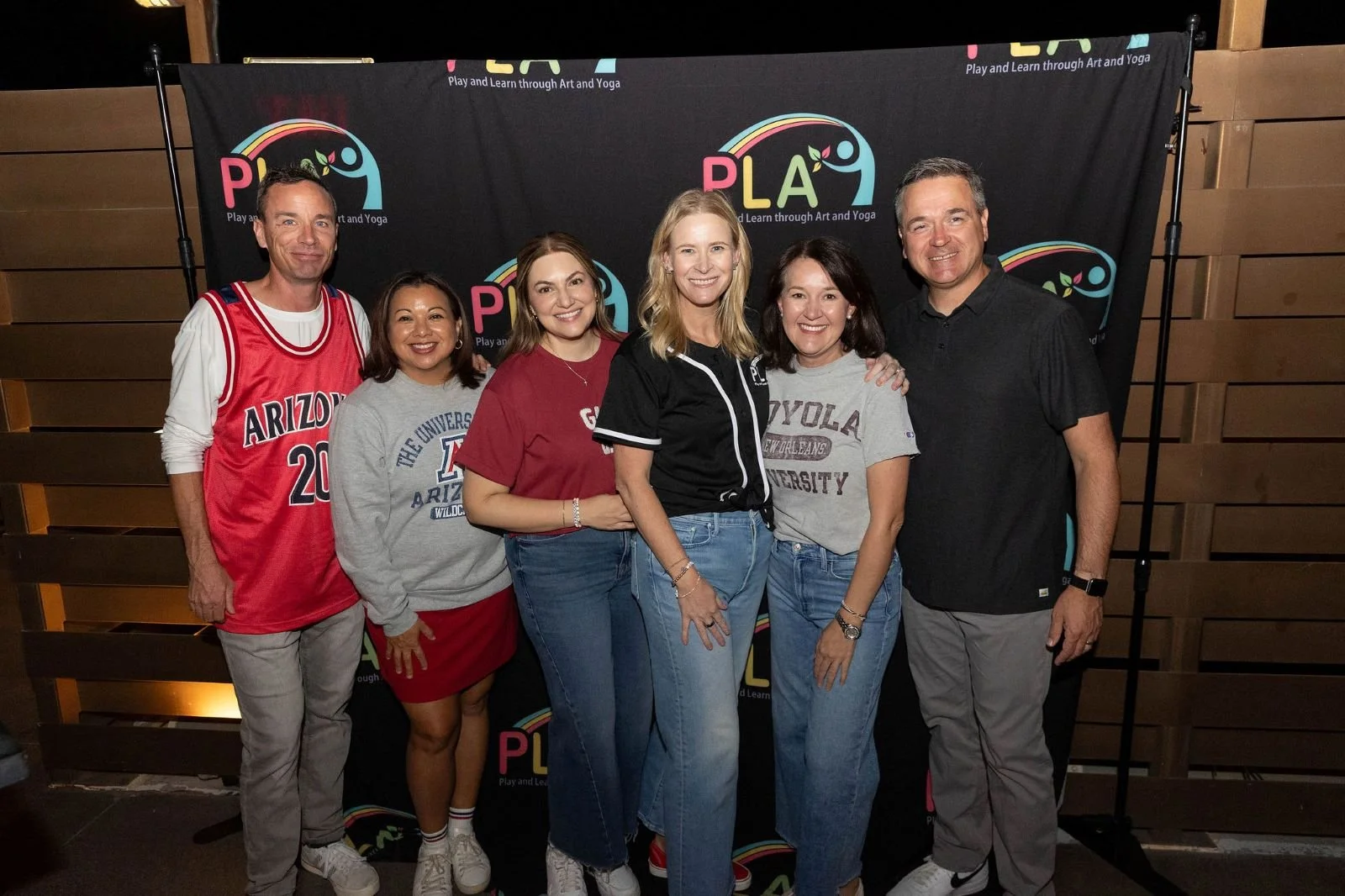 Group of six adults standing together in front of a black backdrop with the Play and Learn through Art and Yoga logo. They are smiling and dressed casually, with some wearing university or team apparel.