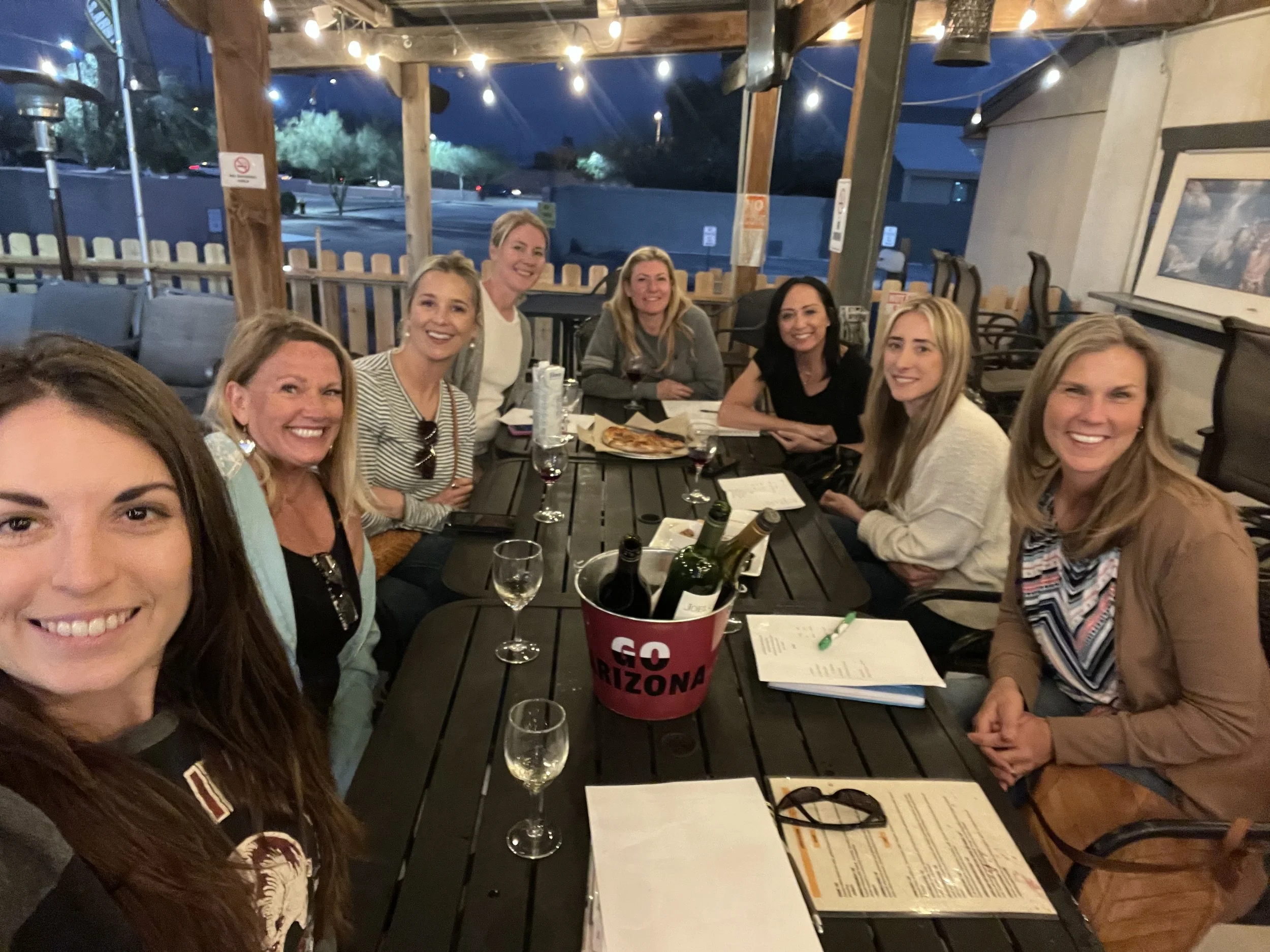 Group of nine women gathered around a table at an outdoor restaurant during evening, enjoying drinks and food.