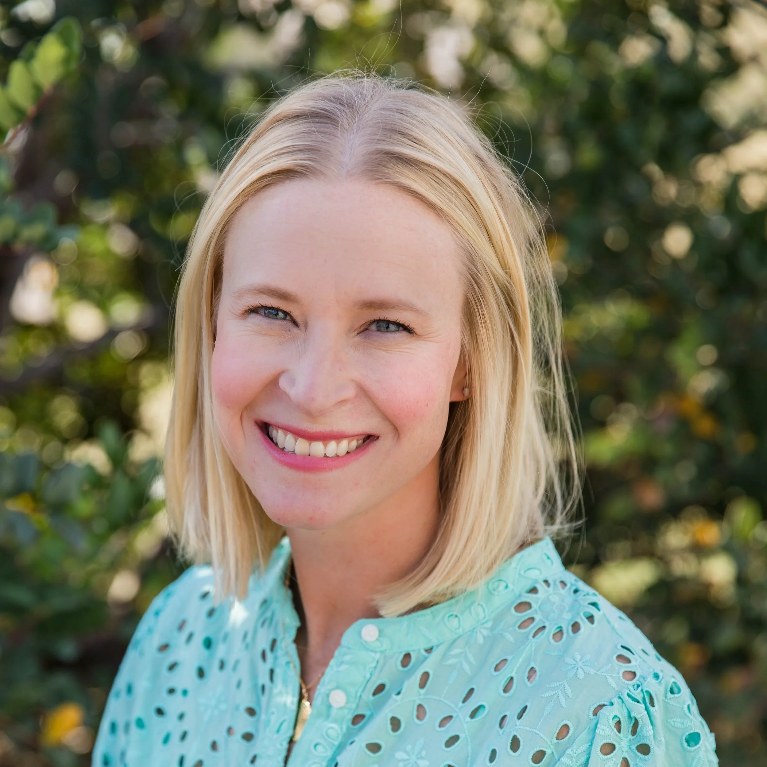 Close-up of a smiling woman with blonde hair and blue eyes, wearing a light blue blouse, outdoors with green foliage in the background.
