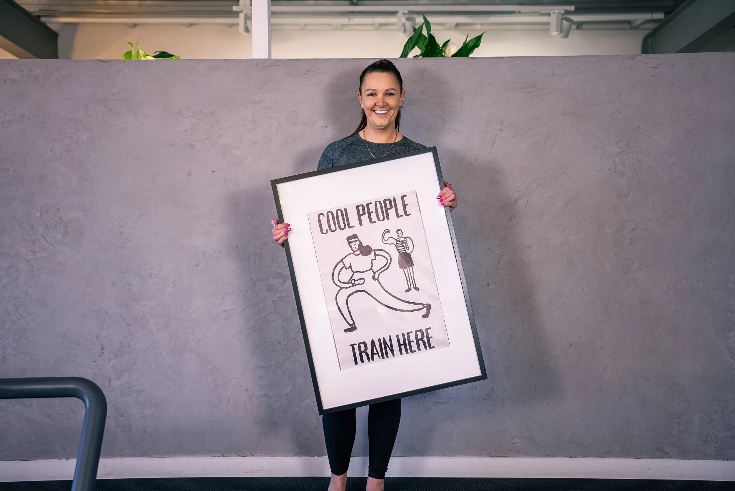 A woman sitting on a Pilates reformer machine at a fitness studio, smiling at the camera, with a wall banner in the background displaying motivational phrases related to Pilates sessions.