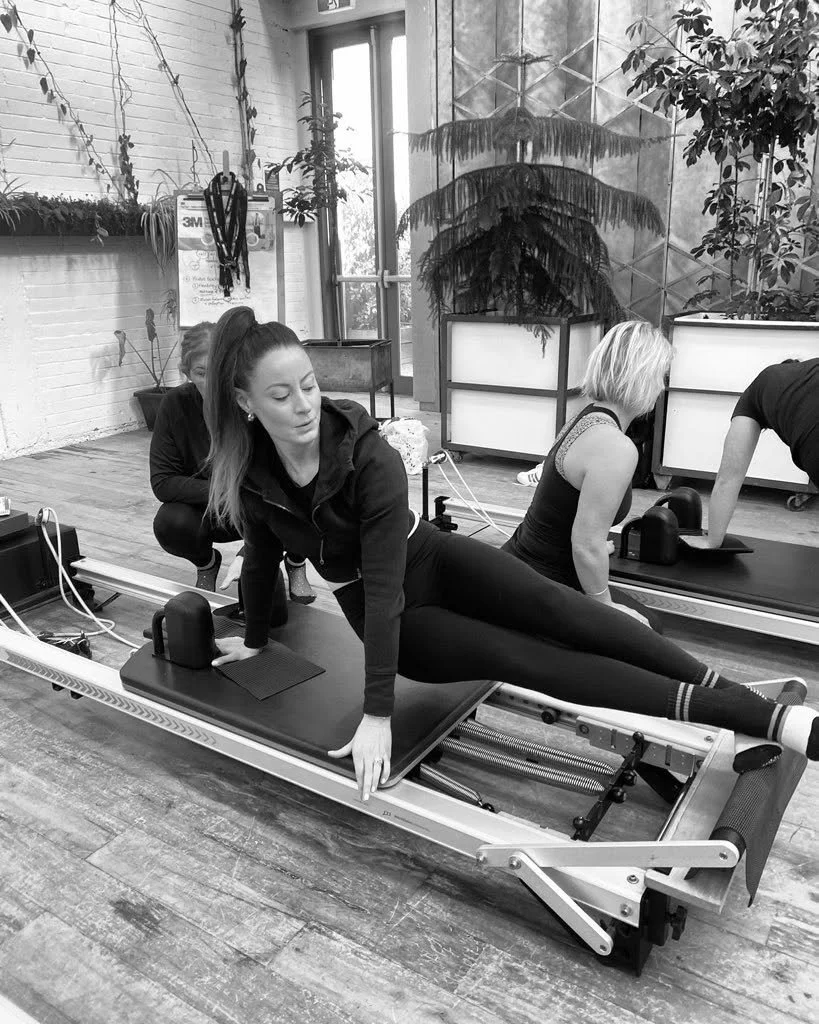 A woman performing a side plank yoga pose on a mat, holding a ball overhead, smiling, in a room with plain wall and ceiling lights.
