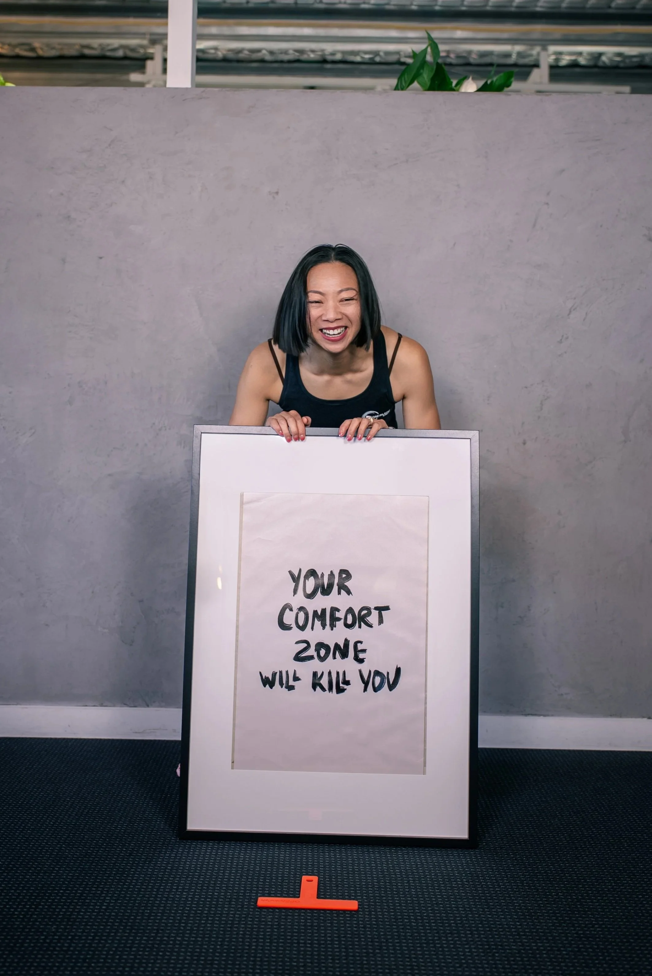 A woman in athletic clothing sitting on a Pilates reformer machine in a fitness studio, with a motivational quote on the wall behind her.