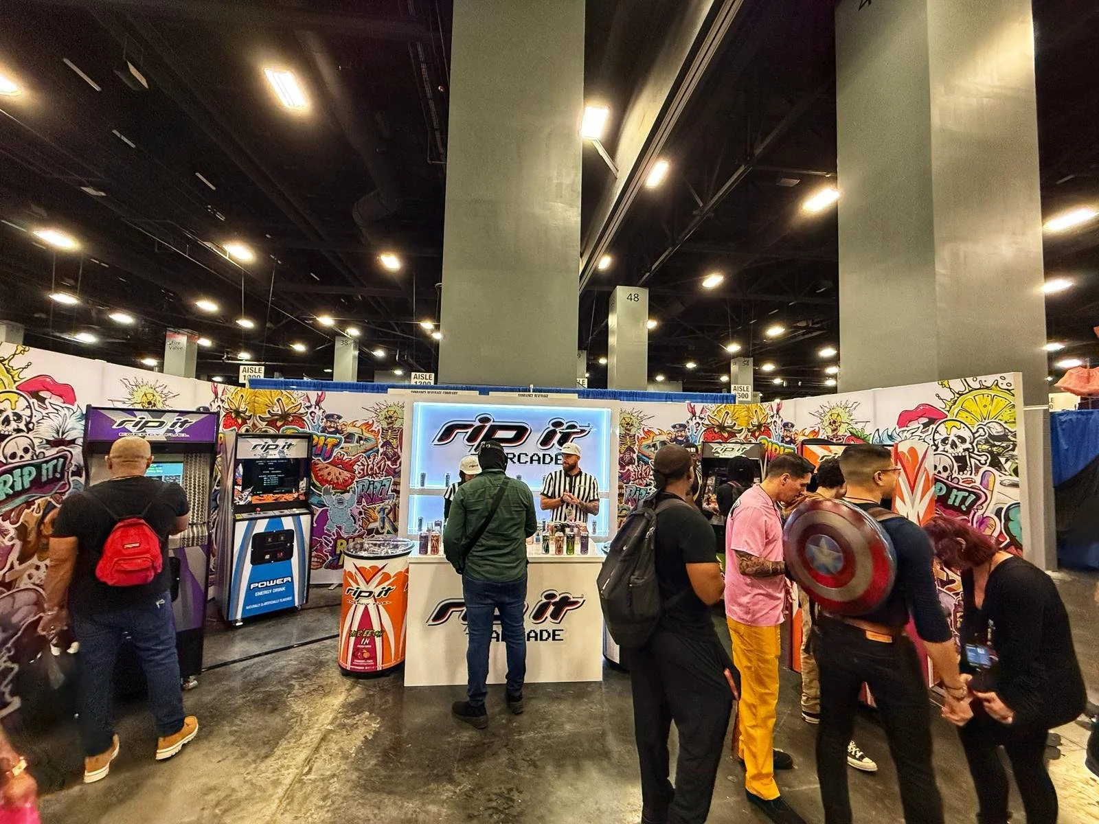 People playing arcade games at Rip It arcade booth, graffiti-style colorful backdrop, indoor convention center setting.