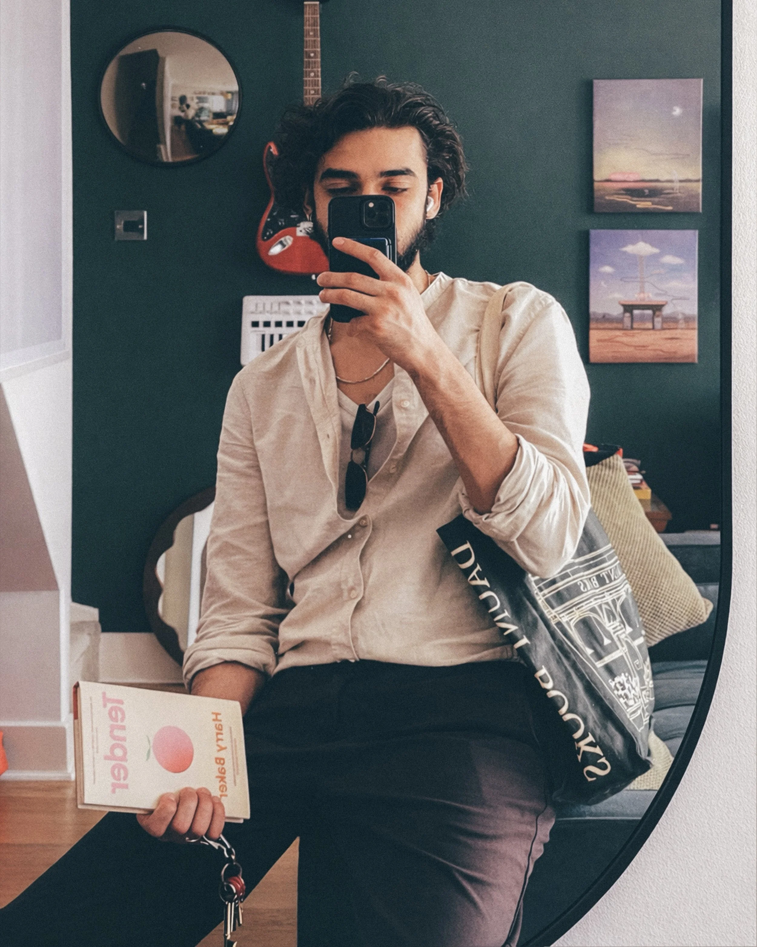 A man taking a mirror selfie in a room with dark green walls, holding a book in his left hand, wearing a beige shirt, black pants, sunglasses hanging from his neckline, and a black bag on his shoulder.