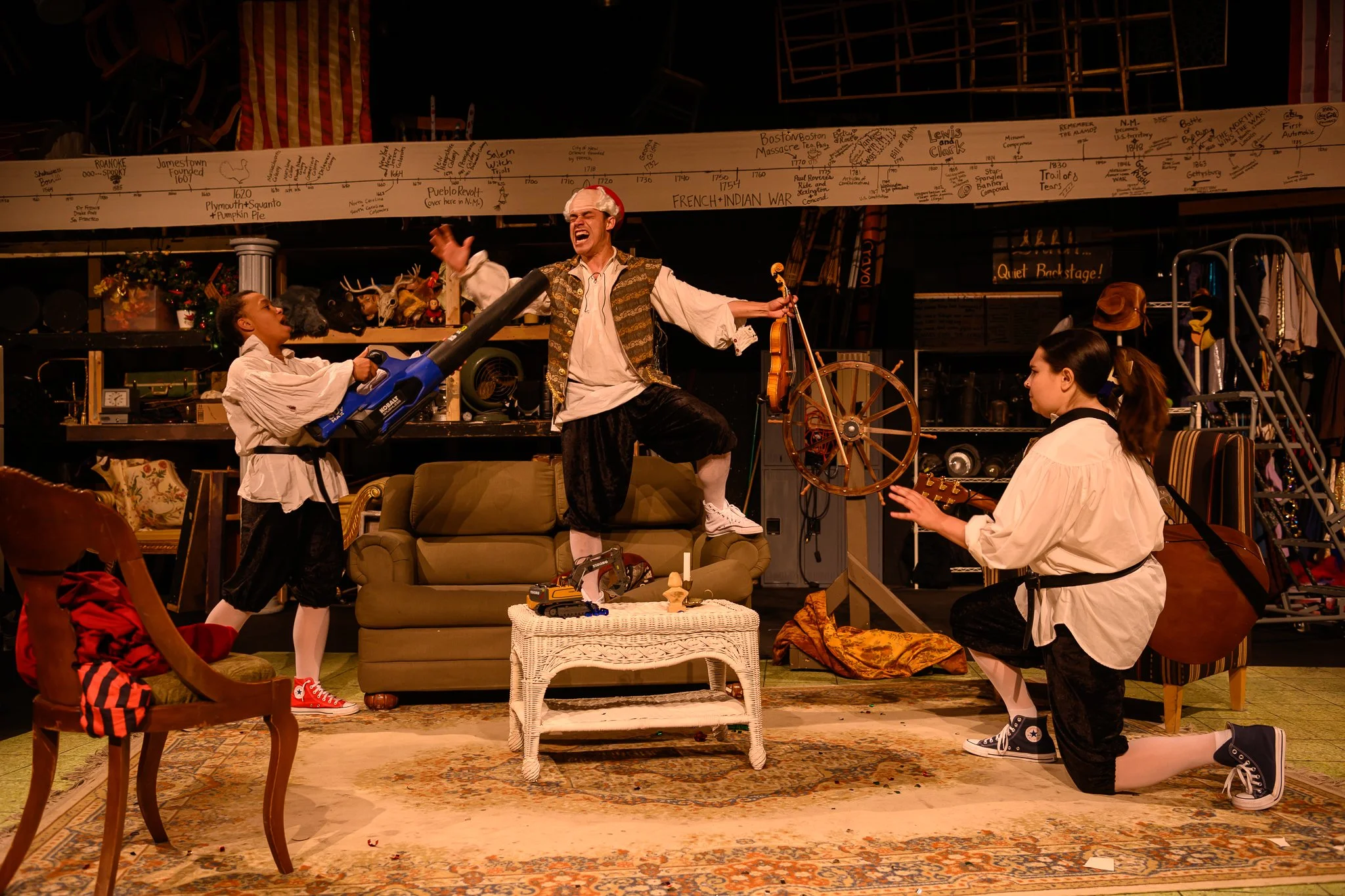 The scene shows a theatrical performance with actors in period costumes. One actor is standing on a couch, mid-action, with expressive gestures. Two other actors are kneeling, engaged with props like a large toy gun and a guitar. The background inclu