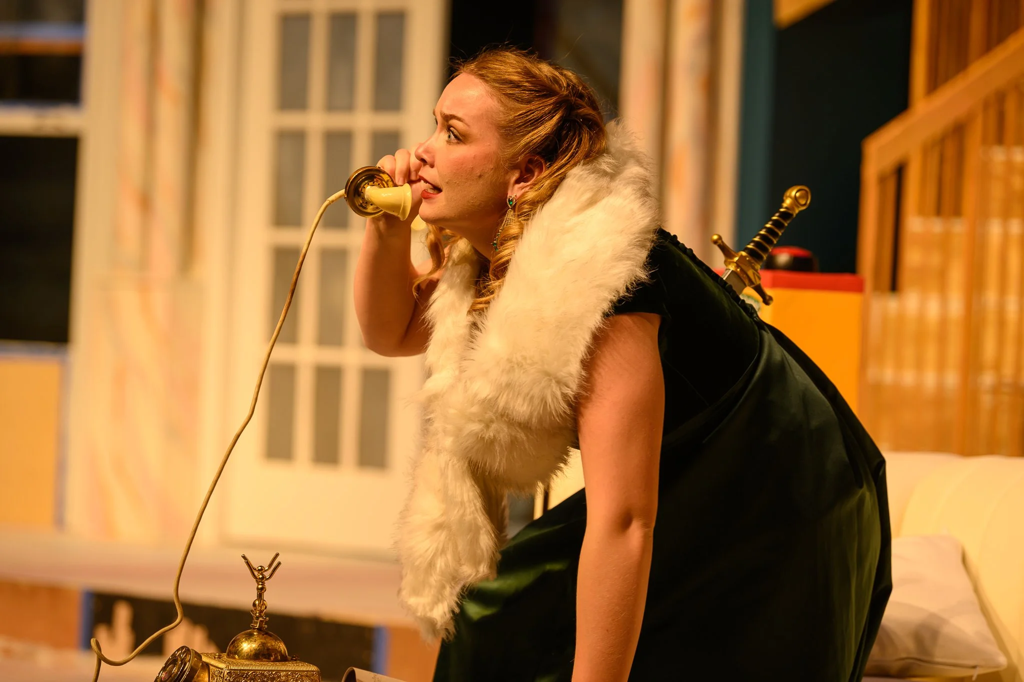 A woman with red hair is on a bed talking on a vintage yellow rotary phone. She has a sword in a sheath on her back, a white fur stole around her shoulders, and is wearing a black dress. The background shows a window and warm lighting.