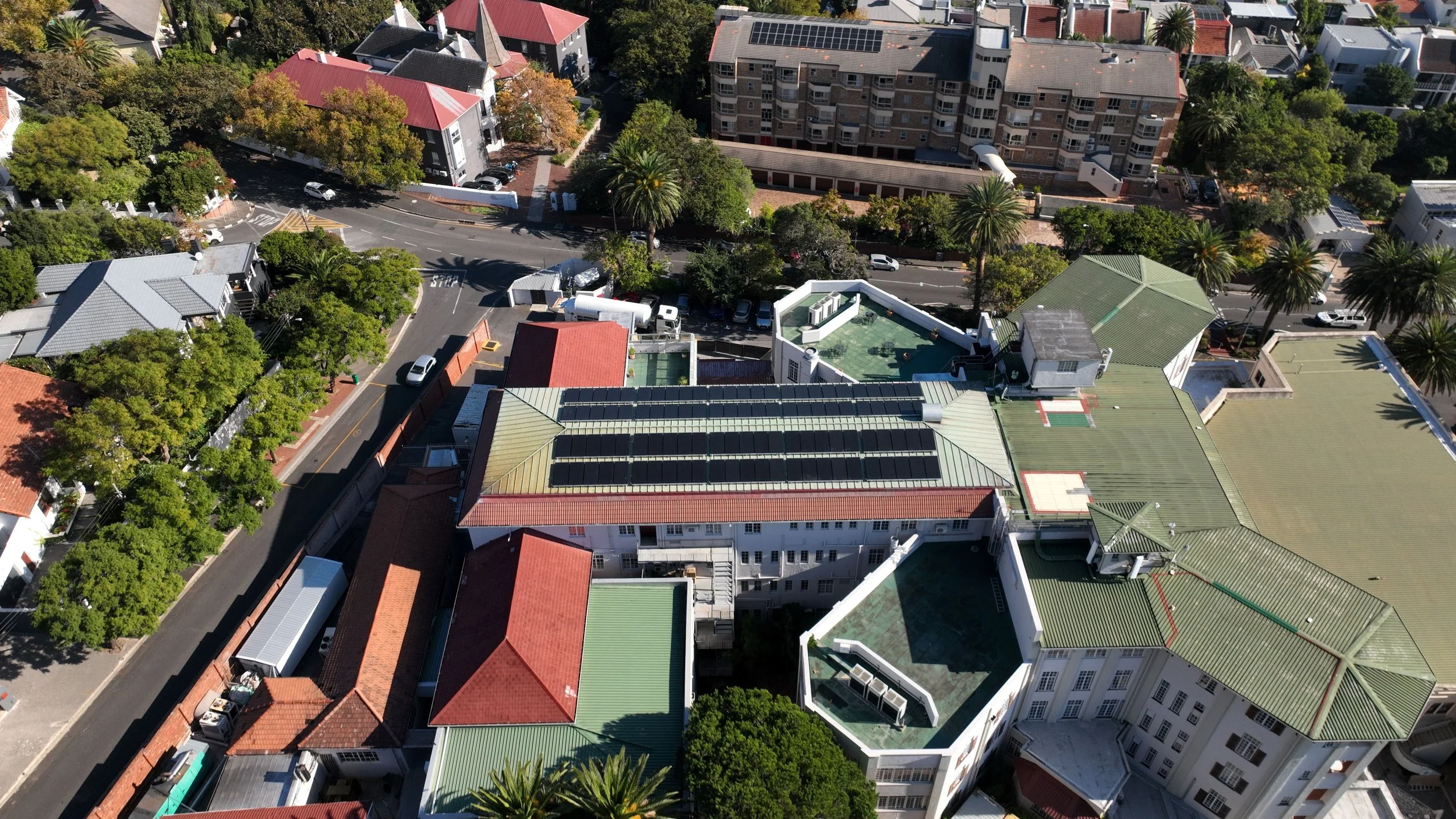 Aerial view of a city block with various buildings, trees, and parking lots. Several rooftops with solar panels, a swimming pool, and streets with cars are visible.