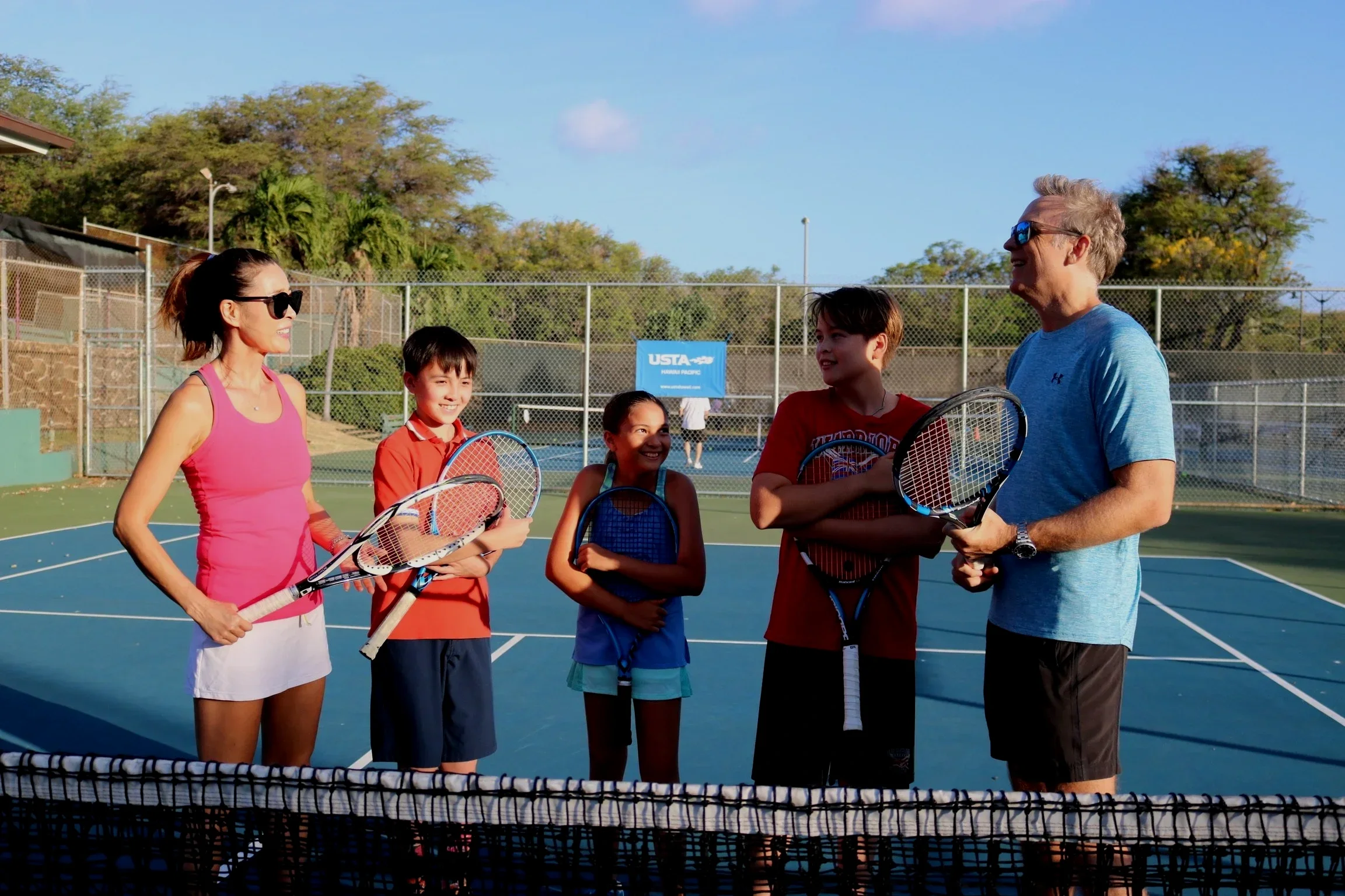 Adult woman and adult man with three children standing on a tennis court, holding tennis rackets, smiling and talking. The court is outdoors with a chain-link fence and a USTA sign in the background, trees, and a clear blue sky.