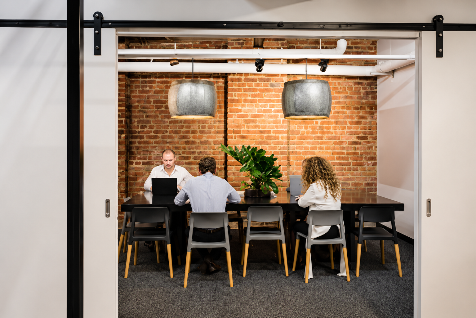 Three people sitting at a black conference table with laptops, in front of a brick wall with two large hanging gray pendant lights and a large potted plant in the middle.