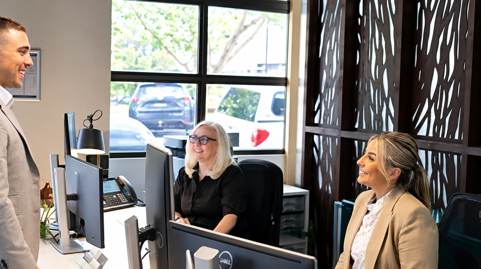 Office reception at rethink your workspace with two women seated at desks and a man standing in front of them, engaged in conversation. The women are smiling, one with blonde hair and glasses, the other with blonde hair in a ponytail.
