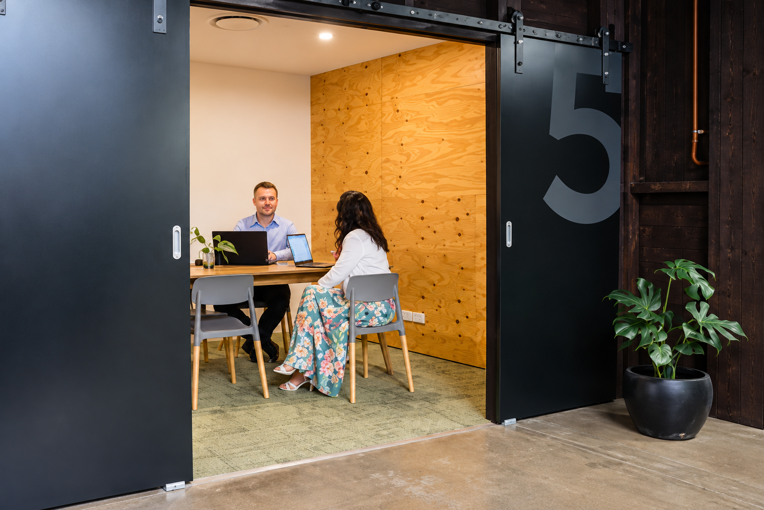 Two people sitting at a table in a modern conference room in newcastle west with wood and dark paneling, seen through a sliding black door with a large number 5, with a potted plant nearby.