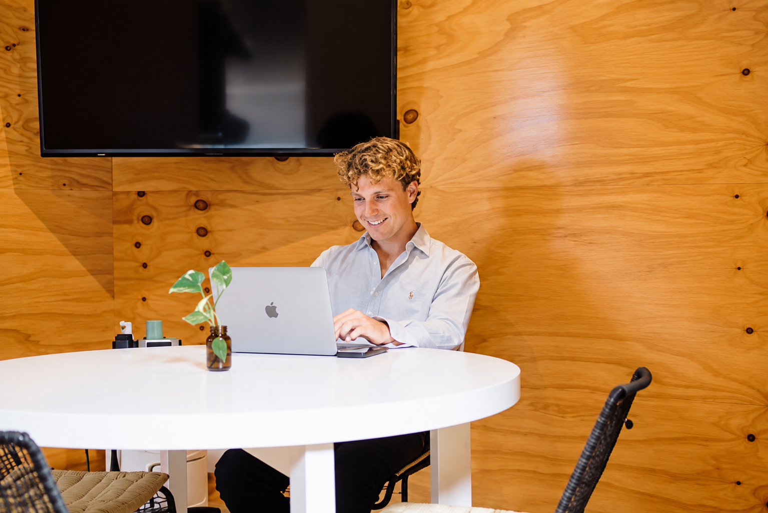 A young man with curly blonde hair sitting at a white round table, working on a silver MacBook, smiling, in a room with wood-paneled walls and a mounted flat-screen TV.