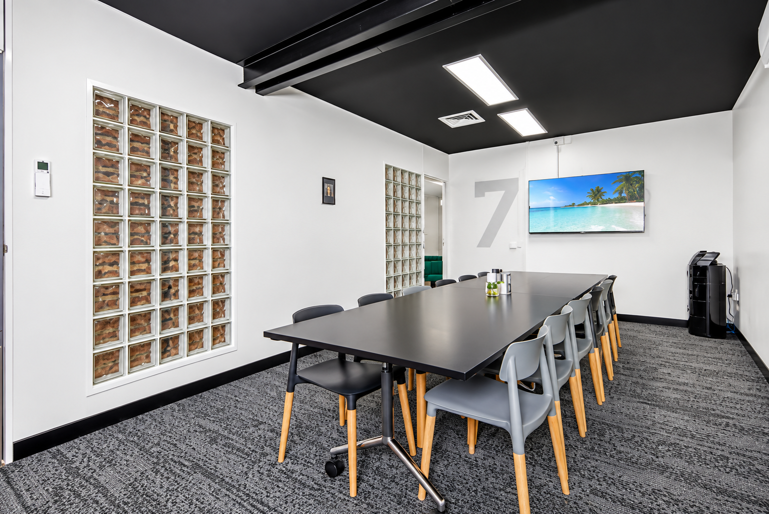 Empty conference room in a newcastle coworking space with black ceiling, white walls, black and gray chairs around a long black table, large TV screen on the wall showing a beach scene, water cooler in corner, and brick wall with glass blocks.