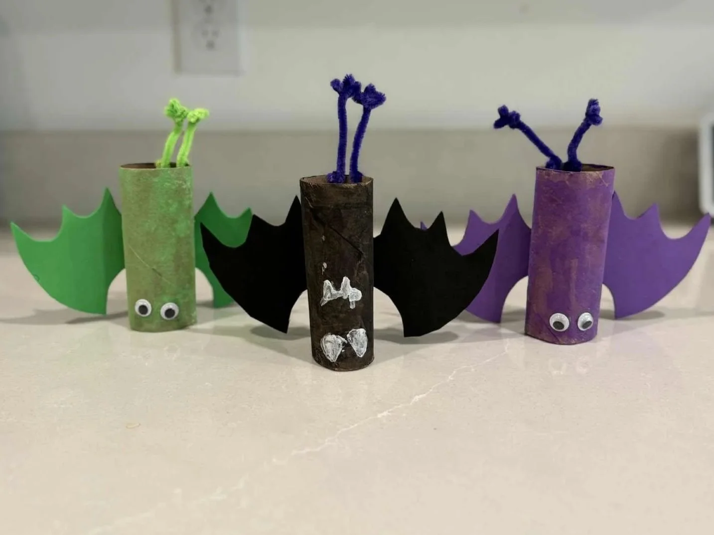 Photo of three bat crafts by kids for Halloween made out of toilet paper rolls and various other craft supplies.