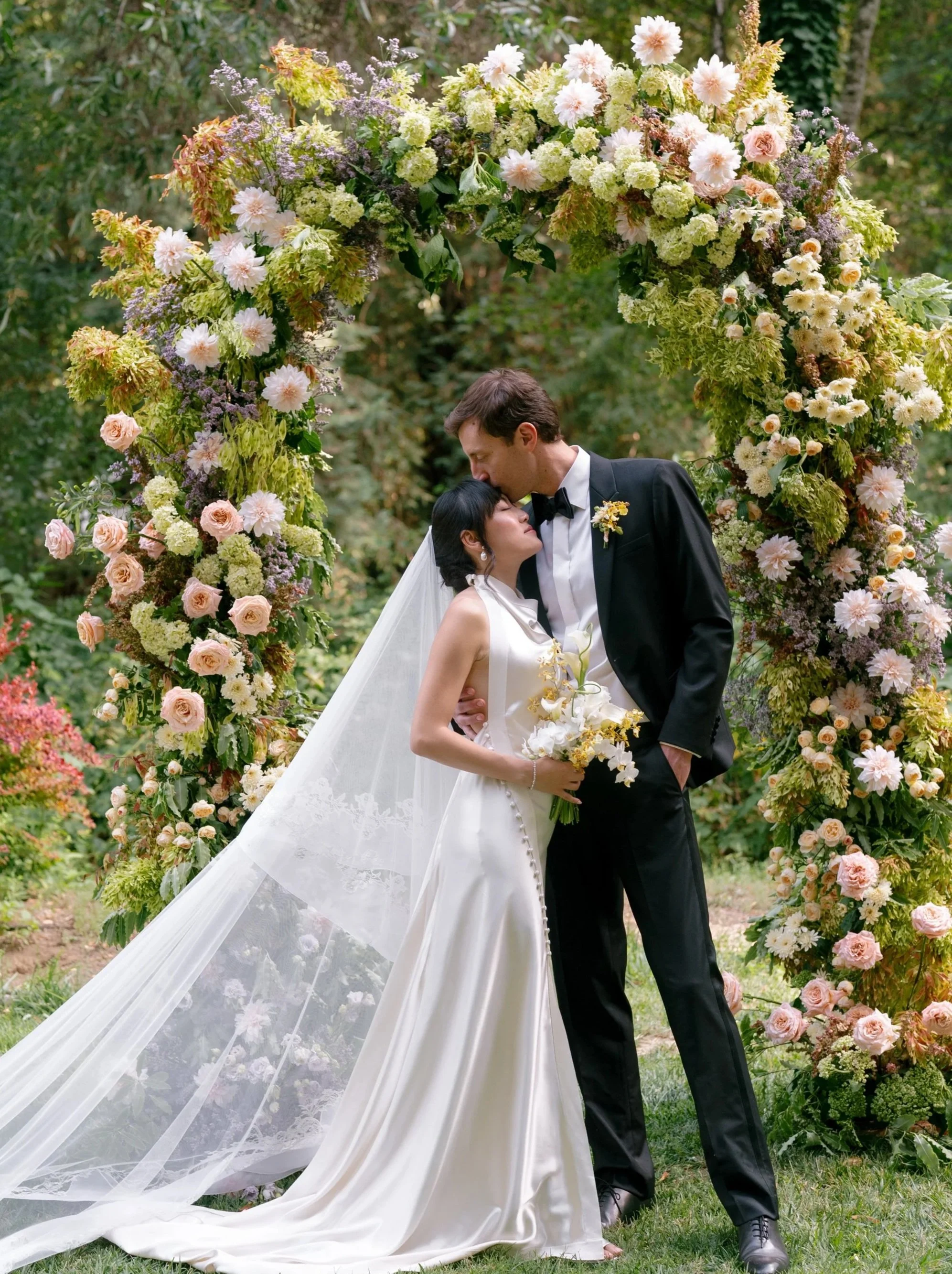 A bride and groom kissing during their outdoor wedding ceremony, standing under a floral arch with pink and white flowers and greenery.