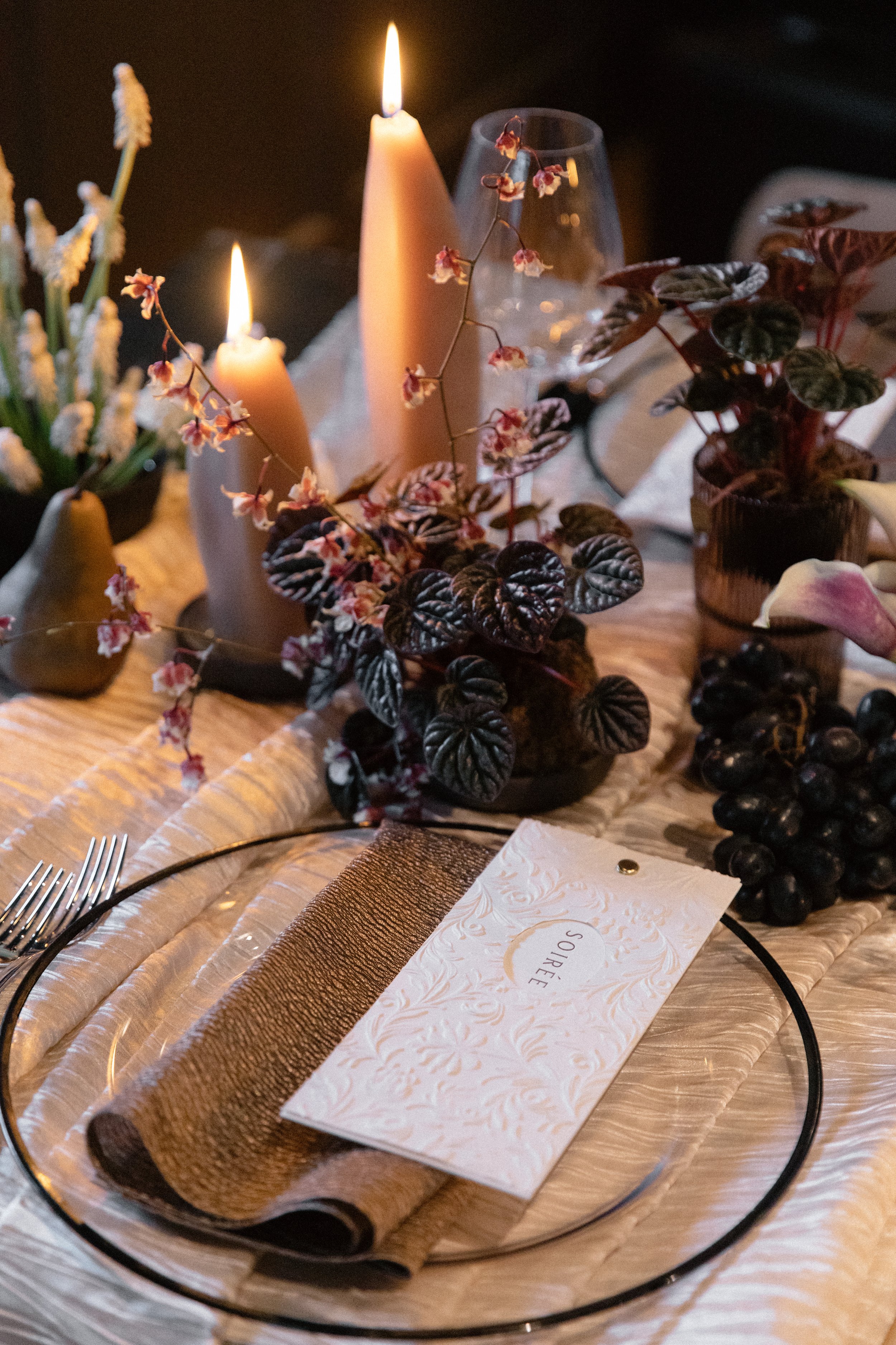 Modern luxury table setting of flowers, plants, candles, and grapes with moody, dim lighting.