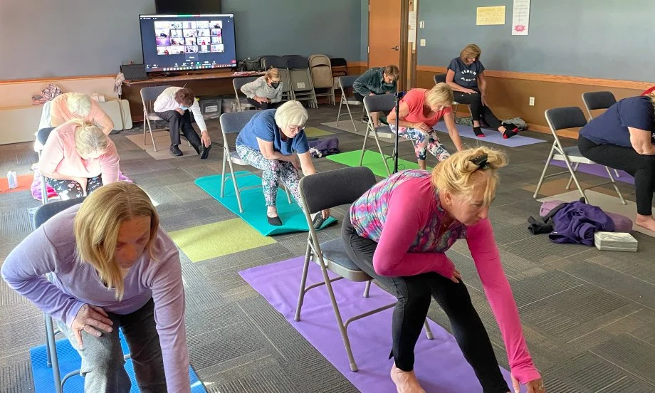 Older adults practicing chair yoga together with online participants visible on a screen.