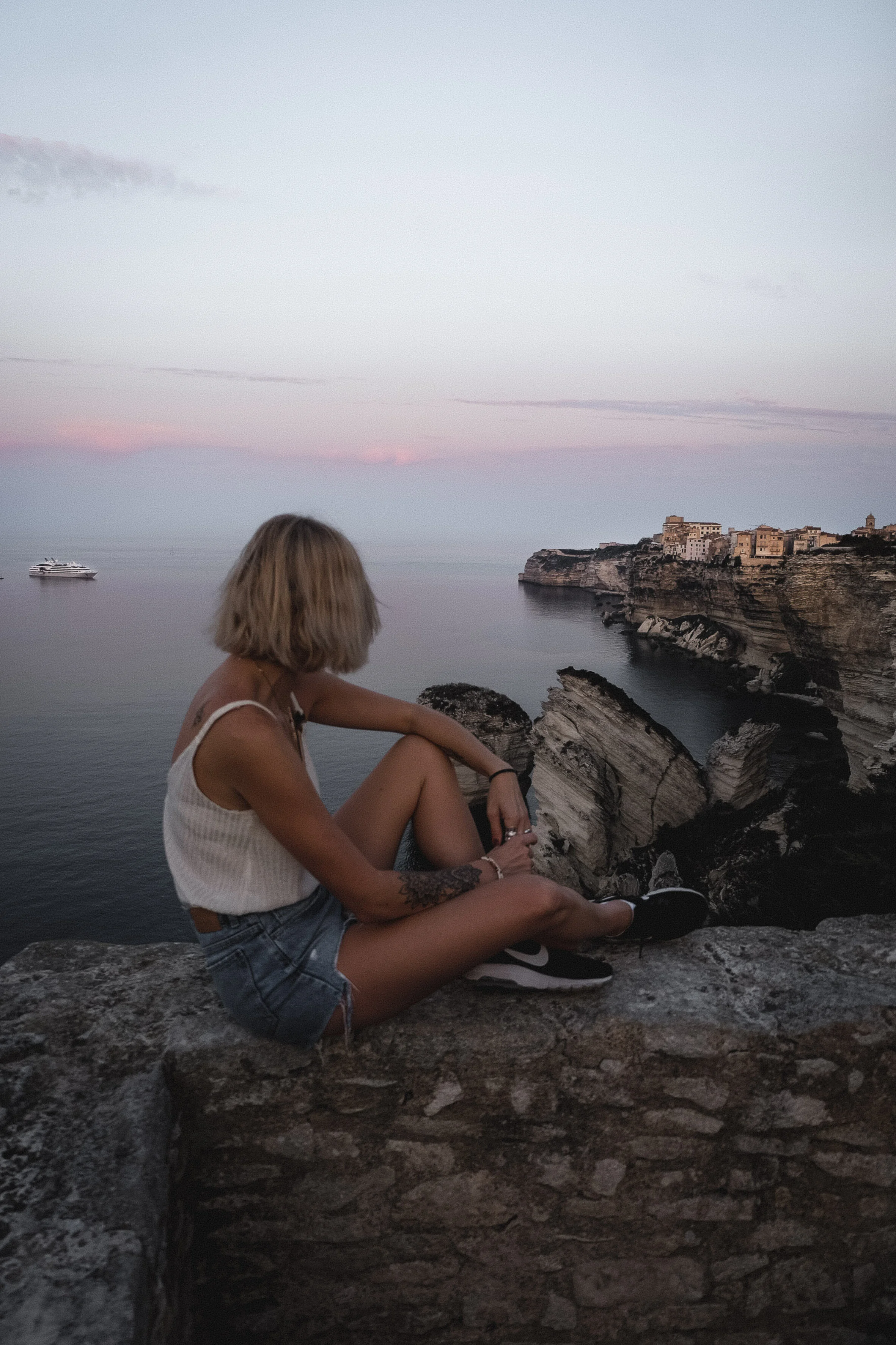 Une femme aux cheveux courts et blonds assise sur un mur en pierre, regardant la mer avec un bateau en arrière-plan et une ville perchée sur une falaise au coucher du soleil.