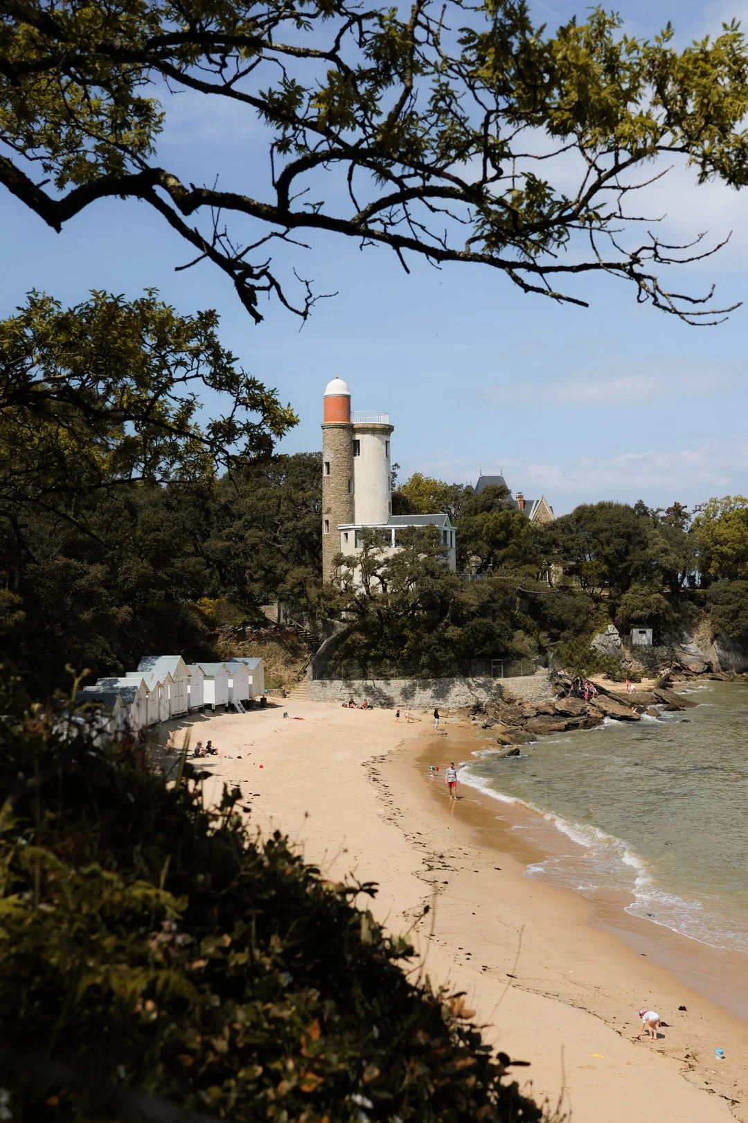 Plage de sable avec des cabines de plage blanches, des personnes qui se baignent et qui se détendent, un phare blanc et rouge situé sur une falaise avec des arbres autour, sous un ciel en partie nuageux.