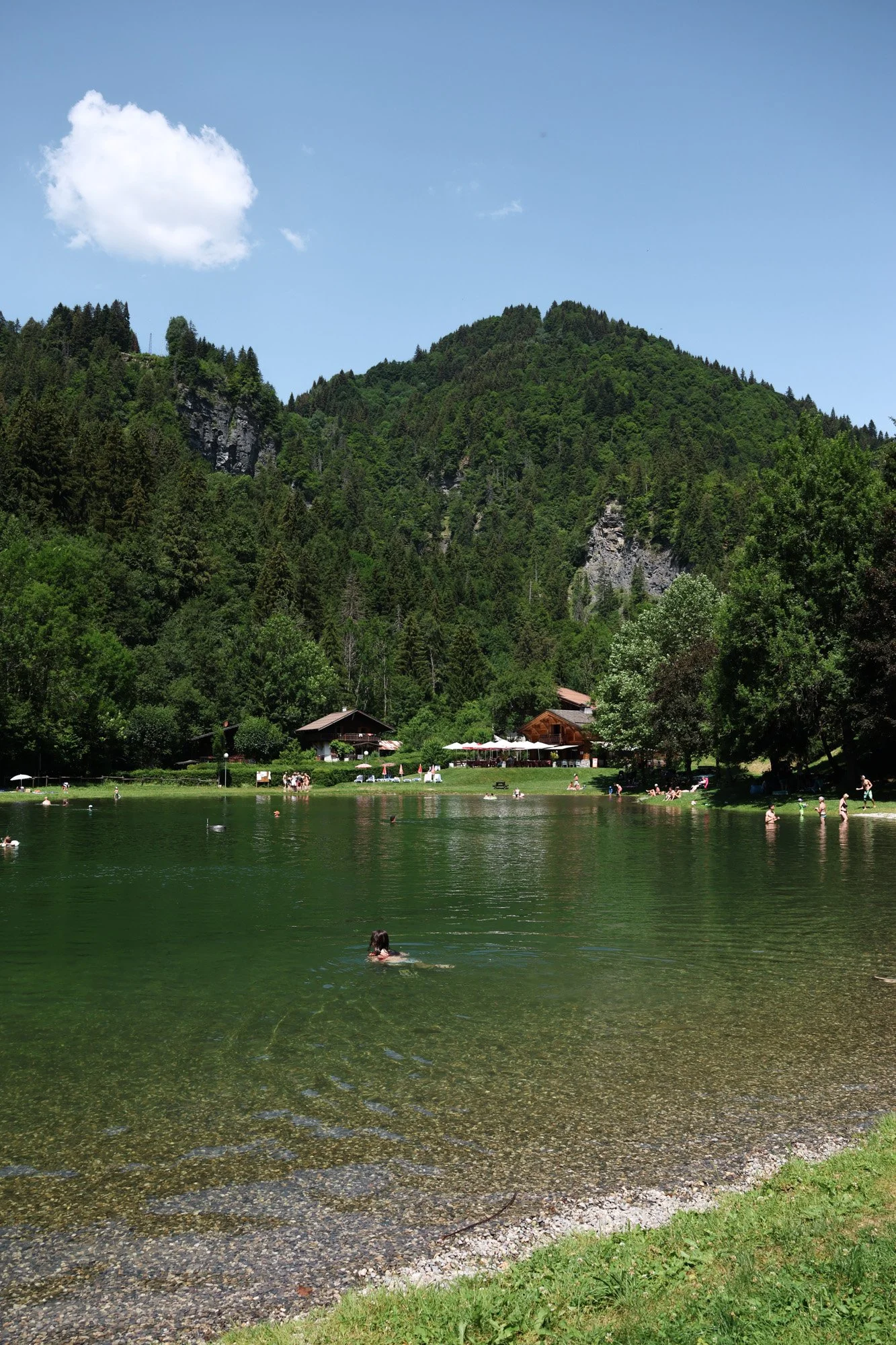 Lac entouré de forêts avec des personnes nageant, des cabanes et une montagne verte en arrière-plan, sous un ciel bleu avec quelques nuages.