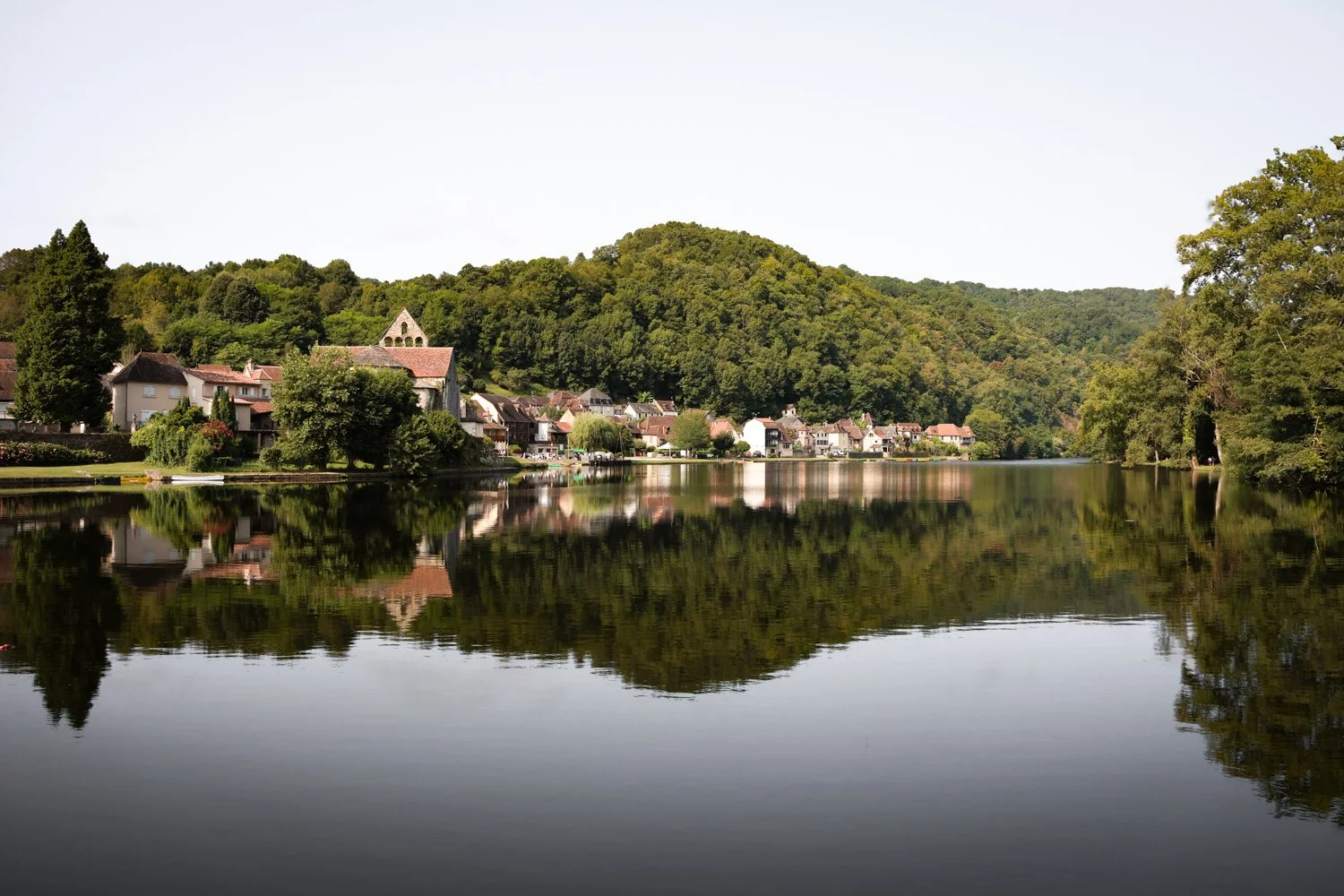 Petit village au bord d'un lac avec maisons, arbres et montagnes en arrière-plan, reflet dans l'eau calme.