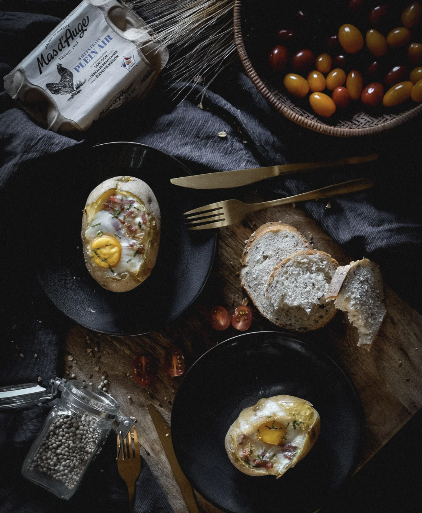 Gros plan sur des toasts garnis d'œufs au plat, de jambon, et d'herbes, accompagnés de tomates miniatures, avec un panier de tomates, un sachet d'œufs, un bol noir, des fourchettes et un moulin à épices blanc, sur une table en bois recouverte d'un ti