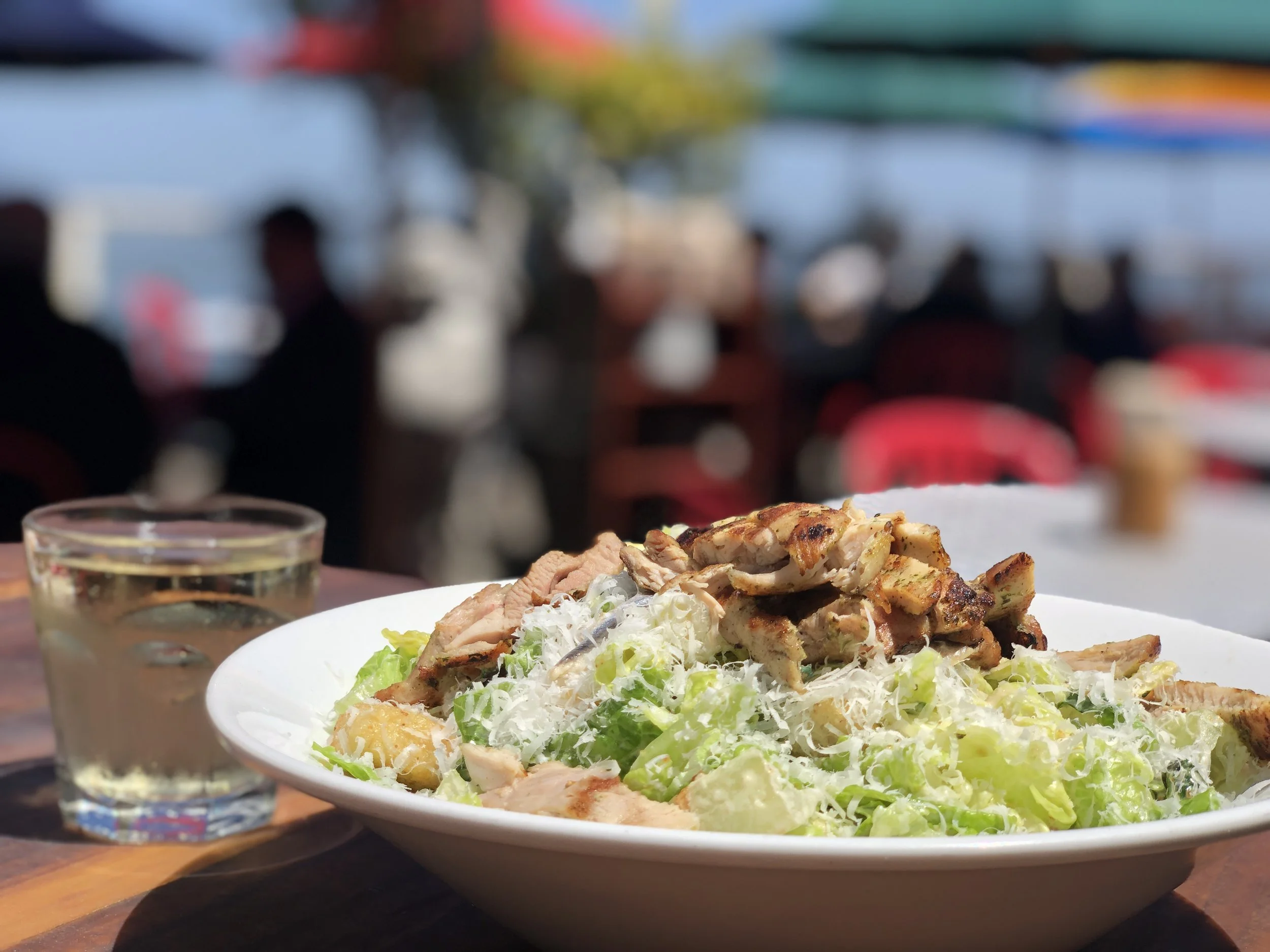 A large white plate with a garden salad topped with grilled chicken and grated cheese, on a wooden table, with a glass of beverage and another plate with food in the background out of focus.