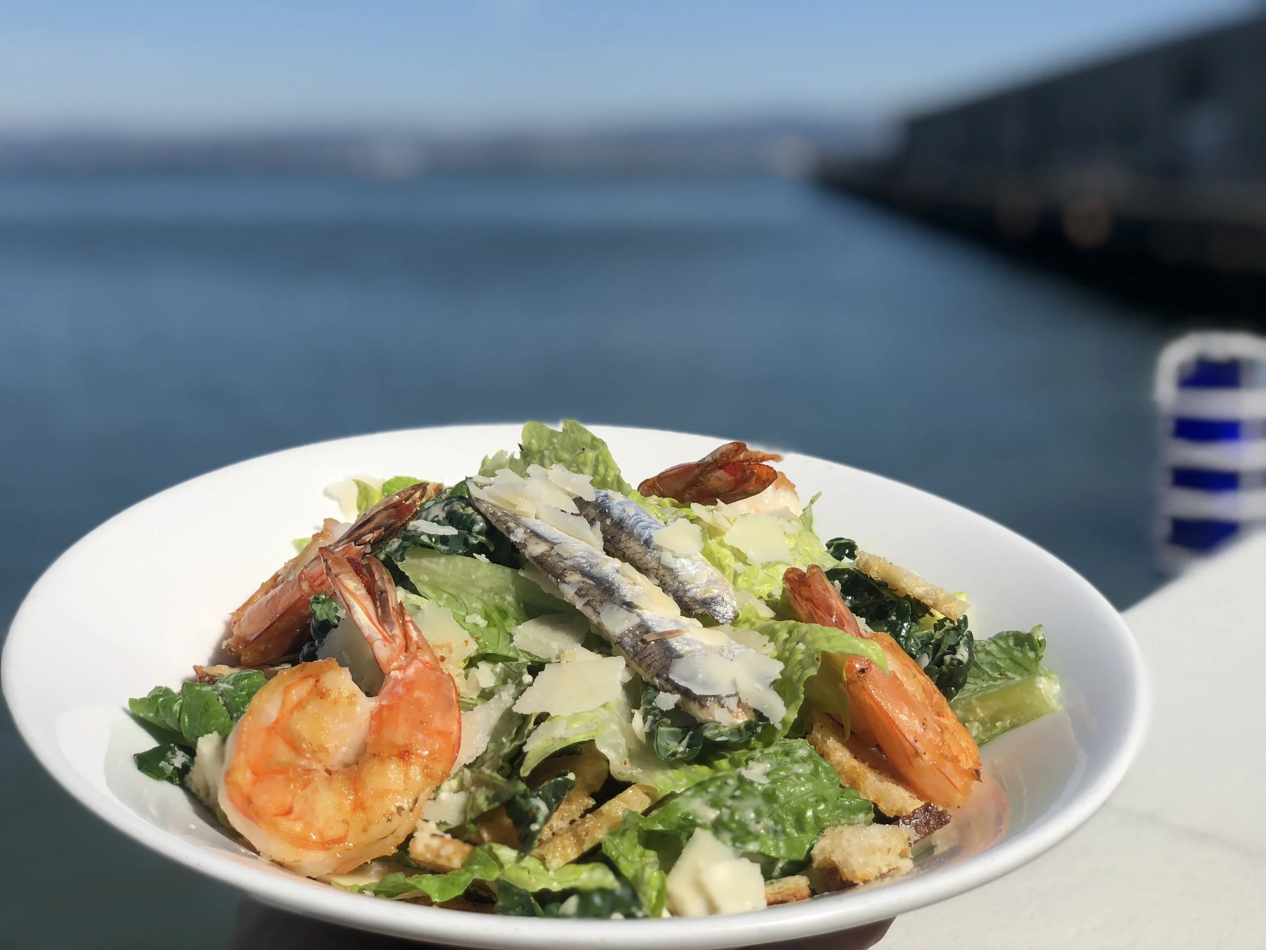 A seafood salad with shrimp, fish, and leafy greens on a white plate outdoors with a blurred ocean background.