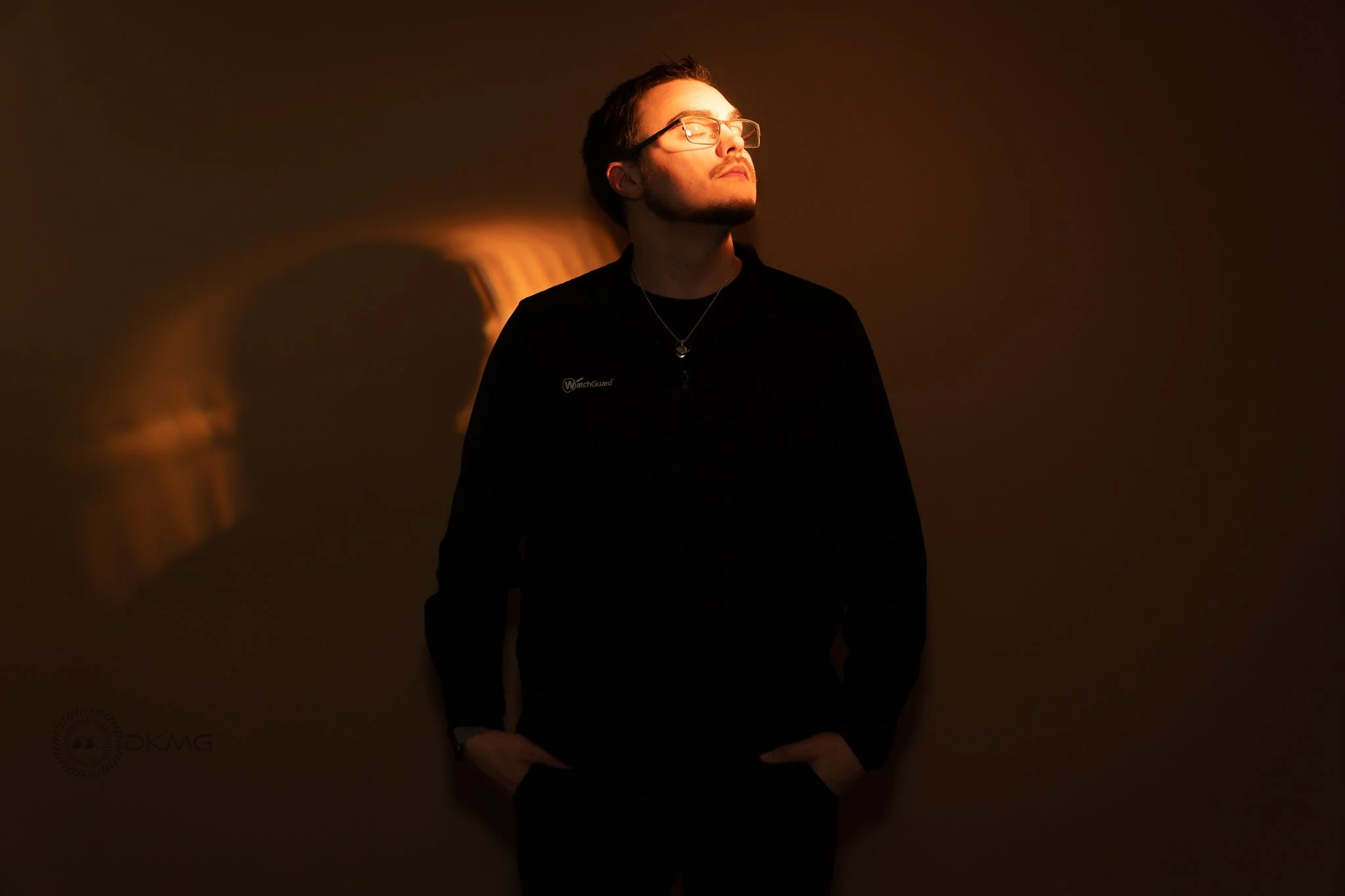 A young man with glasses and a beard standing against a brown wall, with a soft light illuminating part of his face and casting a shadow behind him.