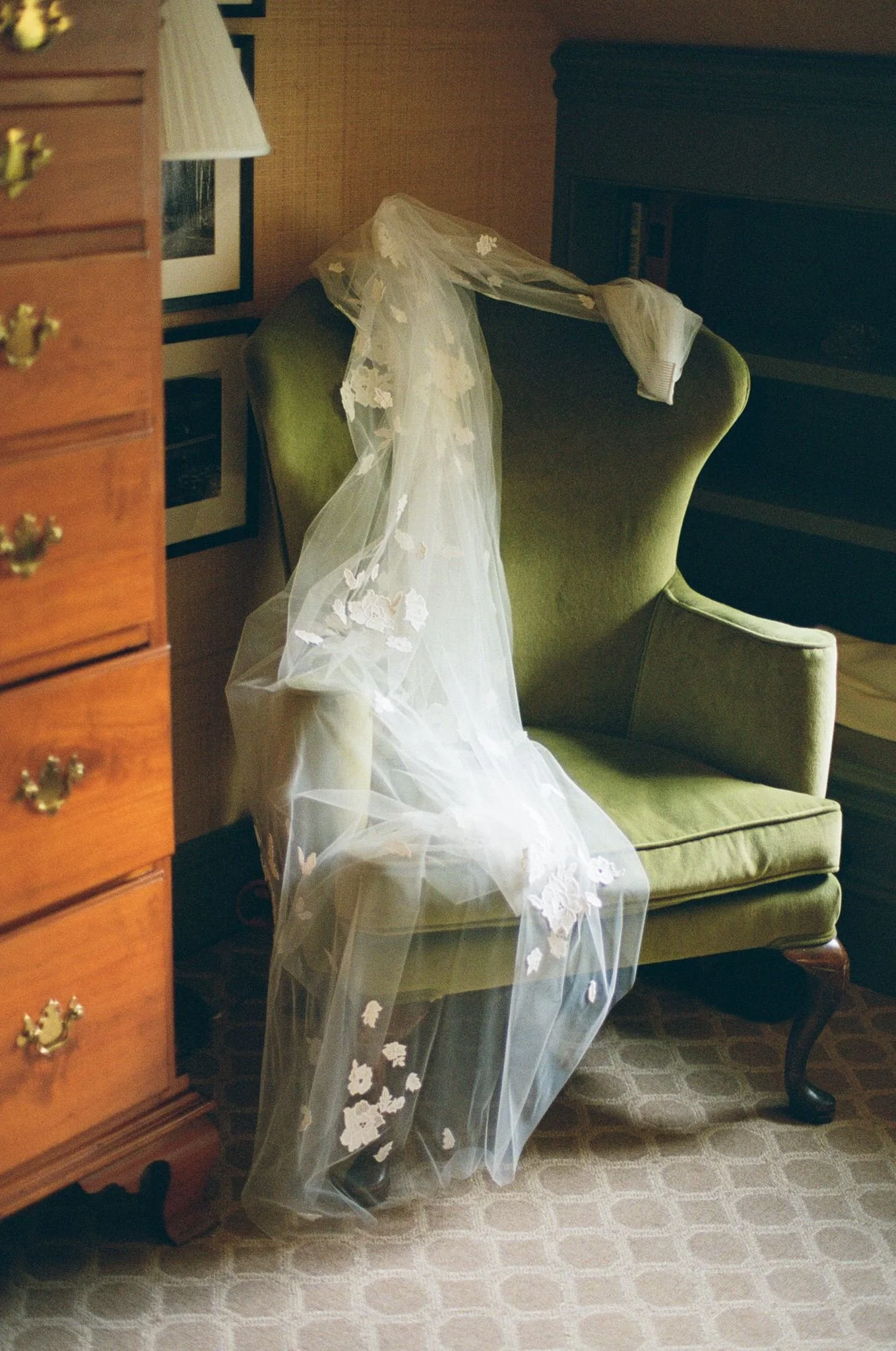 Veil with lace deatosl drapped across a velvet green chair next to a brwon wooden dresser and framed pictures in the background.
